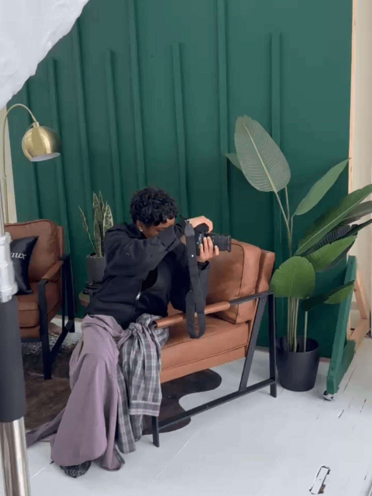 Person sitting on a brown sofa, holding a camera, with large green plants and a green wall behind, in a cozy indoor setting.