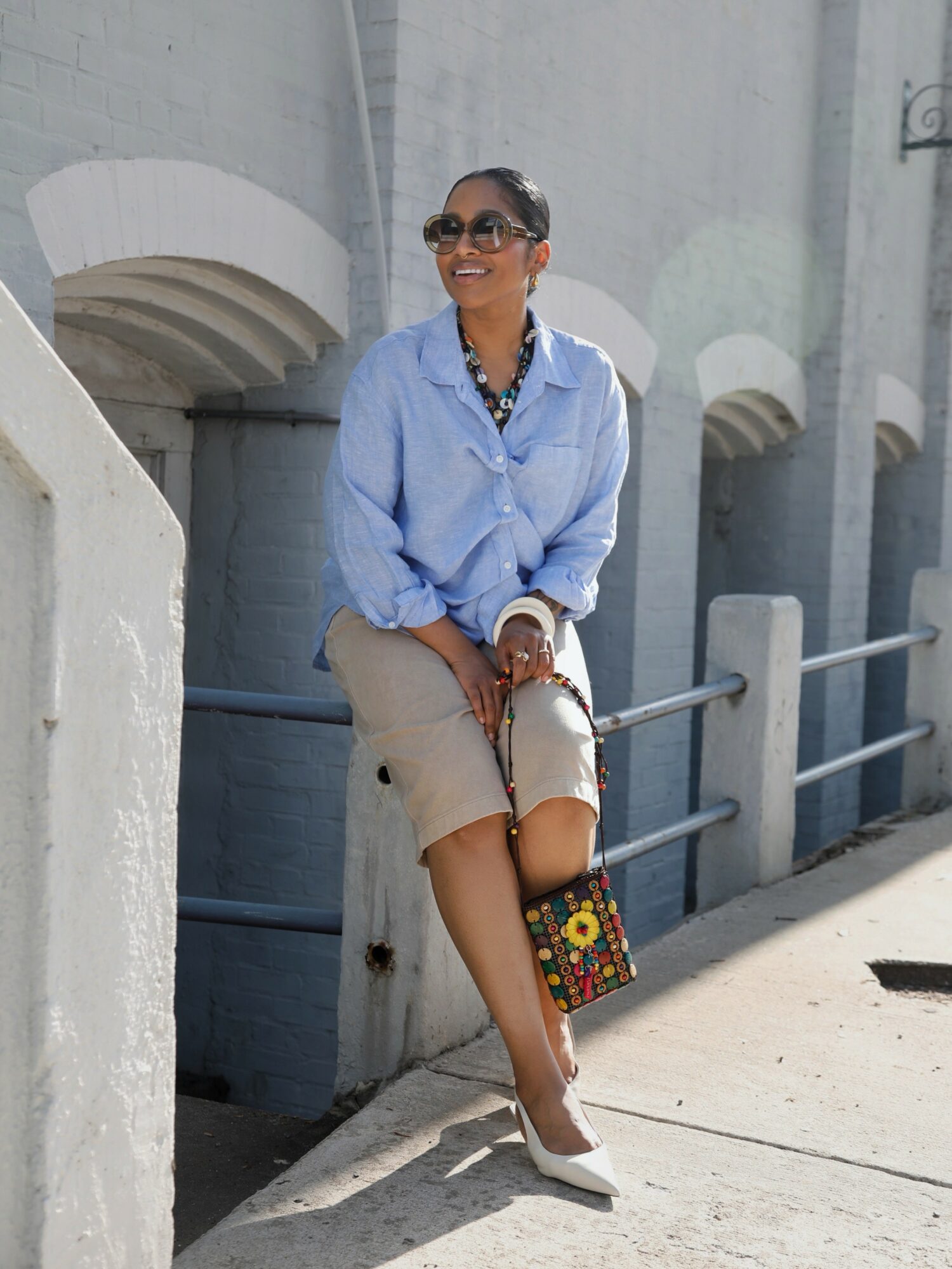 Woman sitting outdoors on a concrete ledge, smiling, wearing sunglasses, a light blue shirt, beige skirt, and white shoes, holding a colorful bag.
