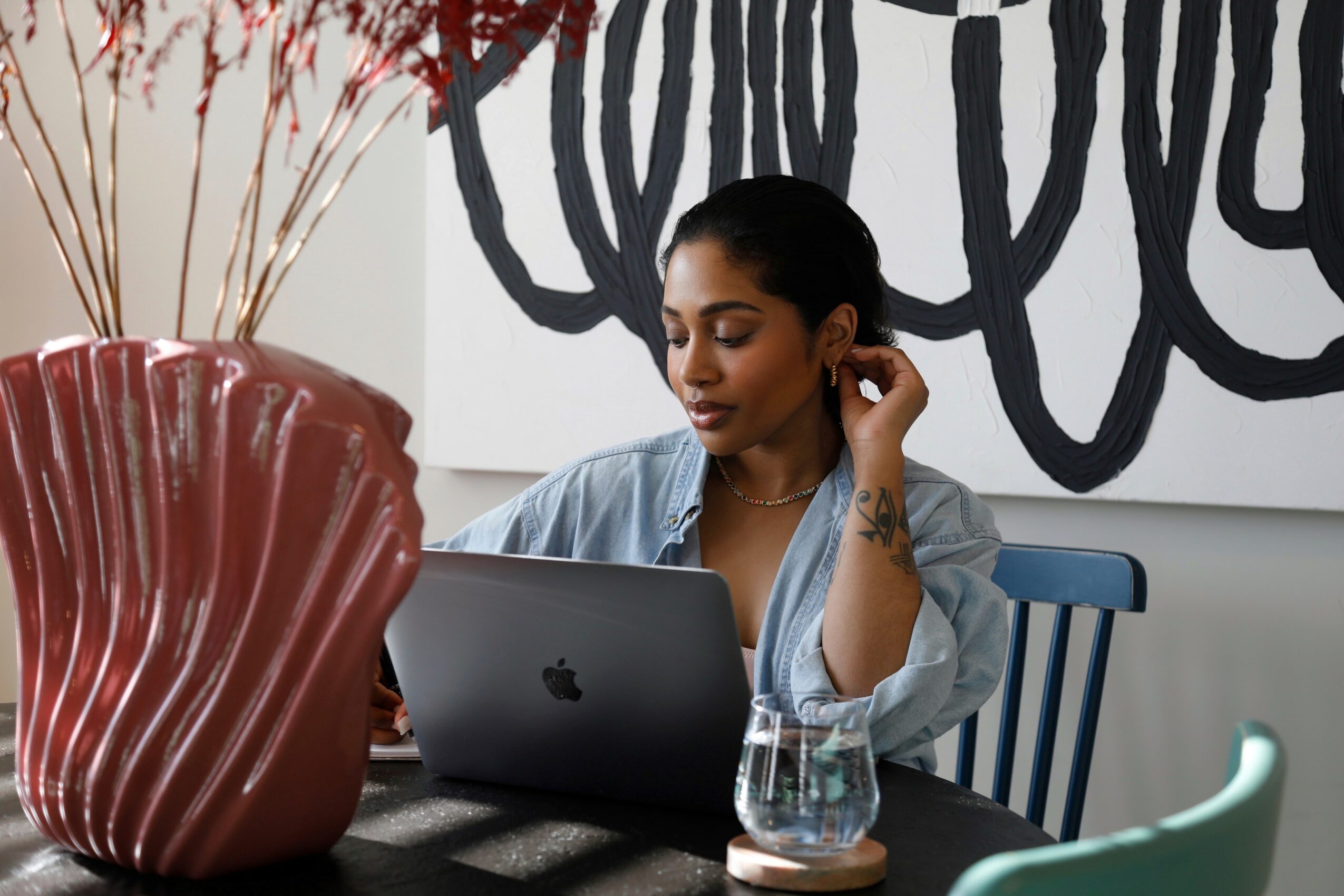 Woman sitting at a table using a laptop, with a large vase and abstract wall art behind her.