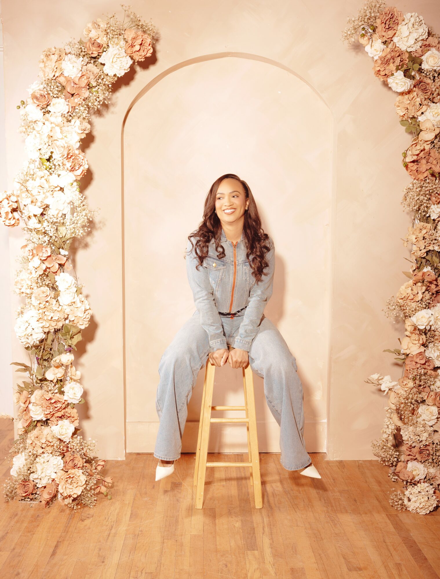 Woman sitting on a wooden stool, smiling, surrounded by floral arch, in a light-colored room with wooden floor.