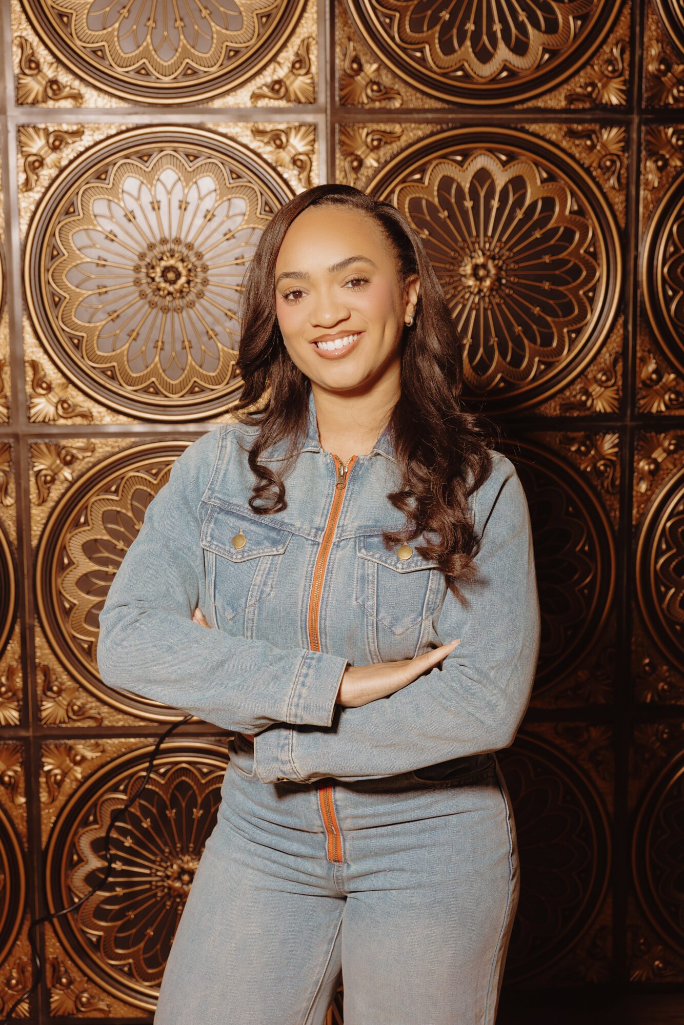 Young woman with long dark hair smiling, standing in front of ornate gold and brown patterned wall, arms crossed, wearing denim outfit.