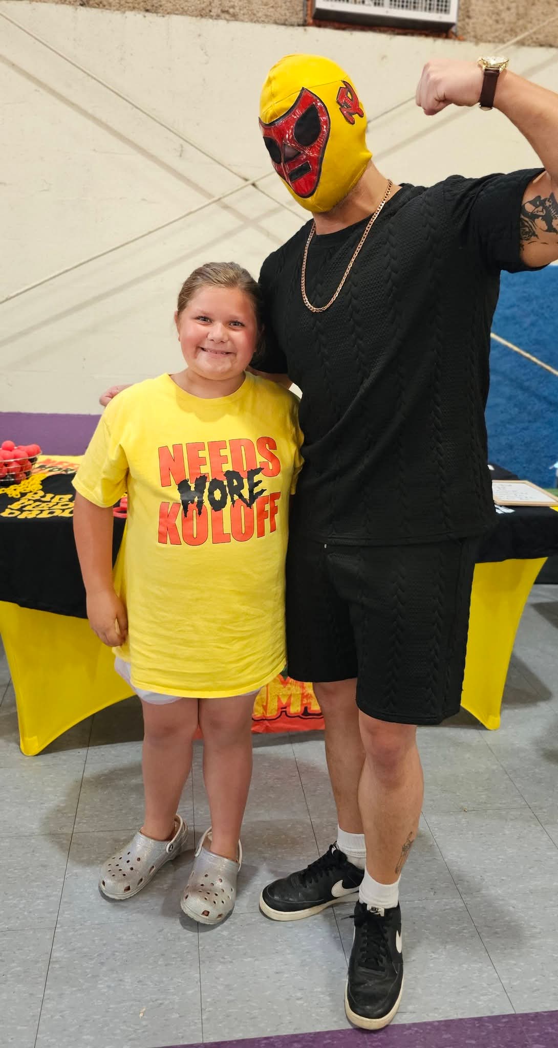 A girl and a person wearing a yellow mask and black clothing pose together indoors, smiling. Girl wears a yellow shirt with red text.