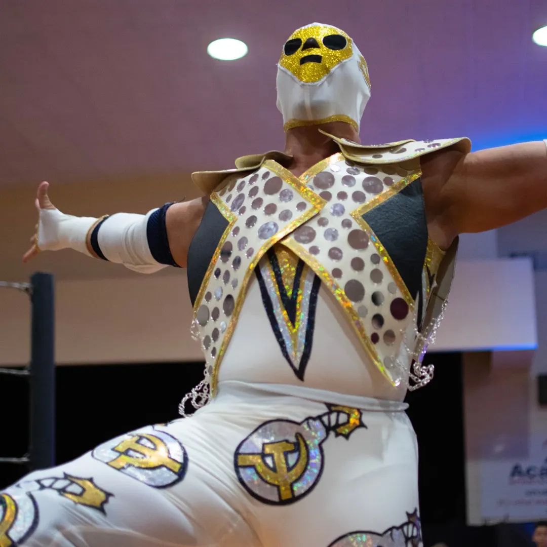 Person in colorful costume with a mask, arms outstretched, indoors with ceiling lights visible.