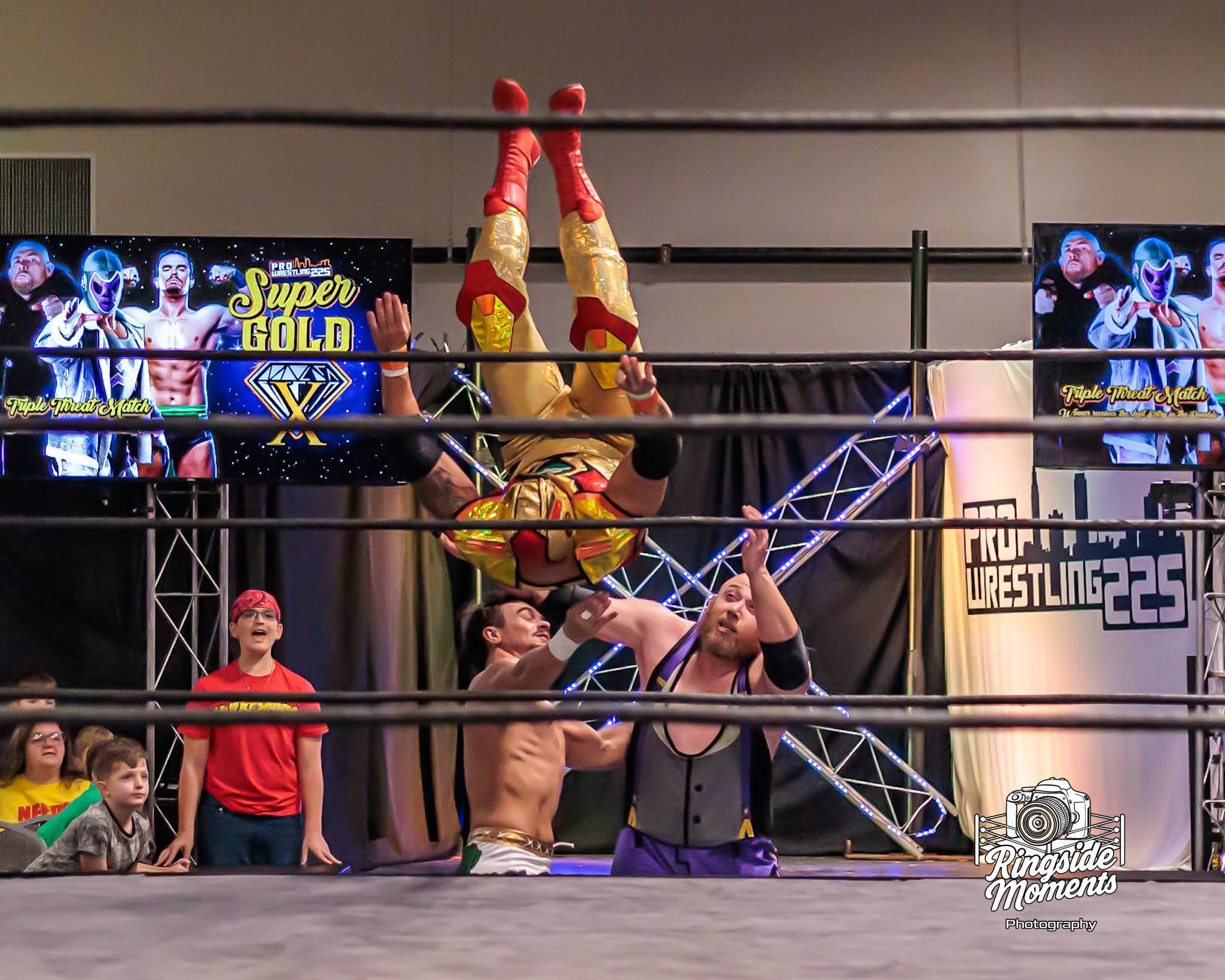 Two wrestlers perform a move with one upside down, others and spectators watch inside a wrestling ring. Bright screens display graphics.