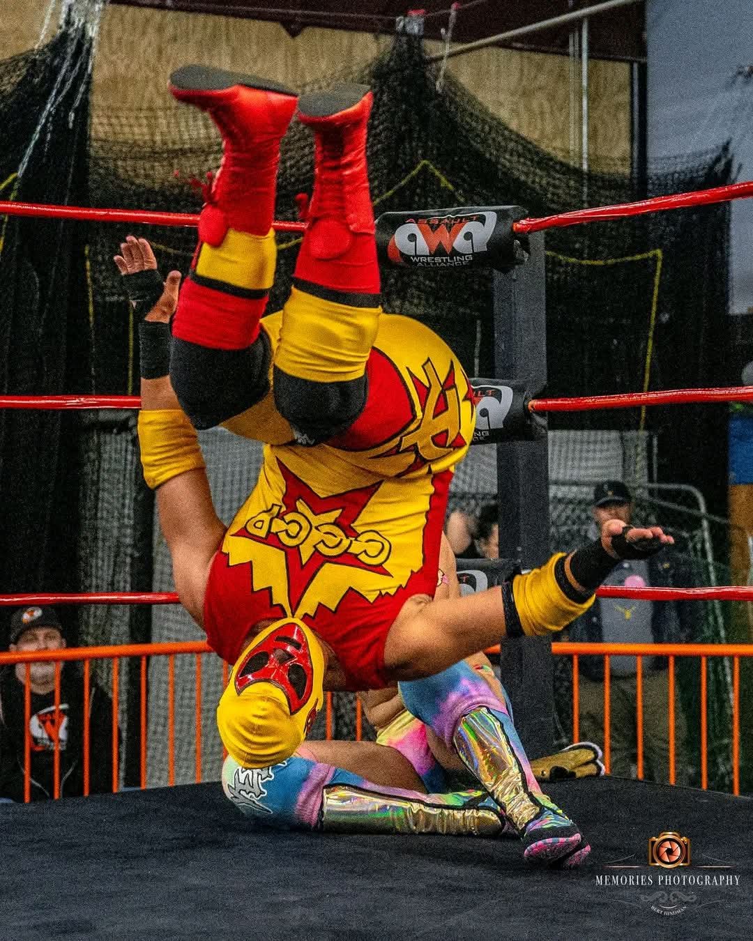 Wrestler in red and yellow costume upside down in wrestling ring, with another wrestler on the ground.