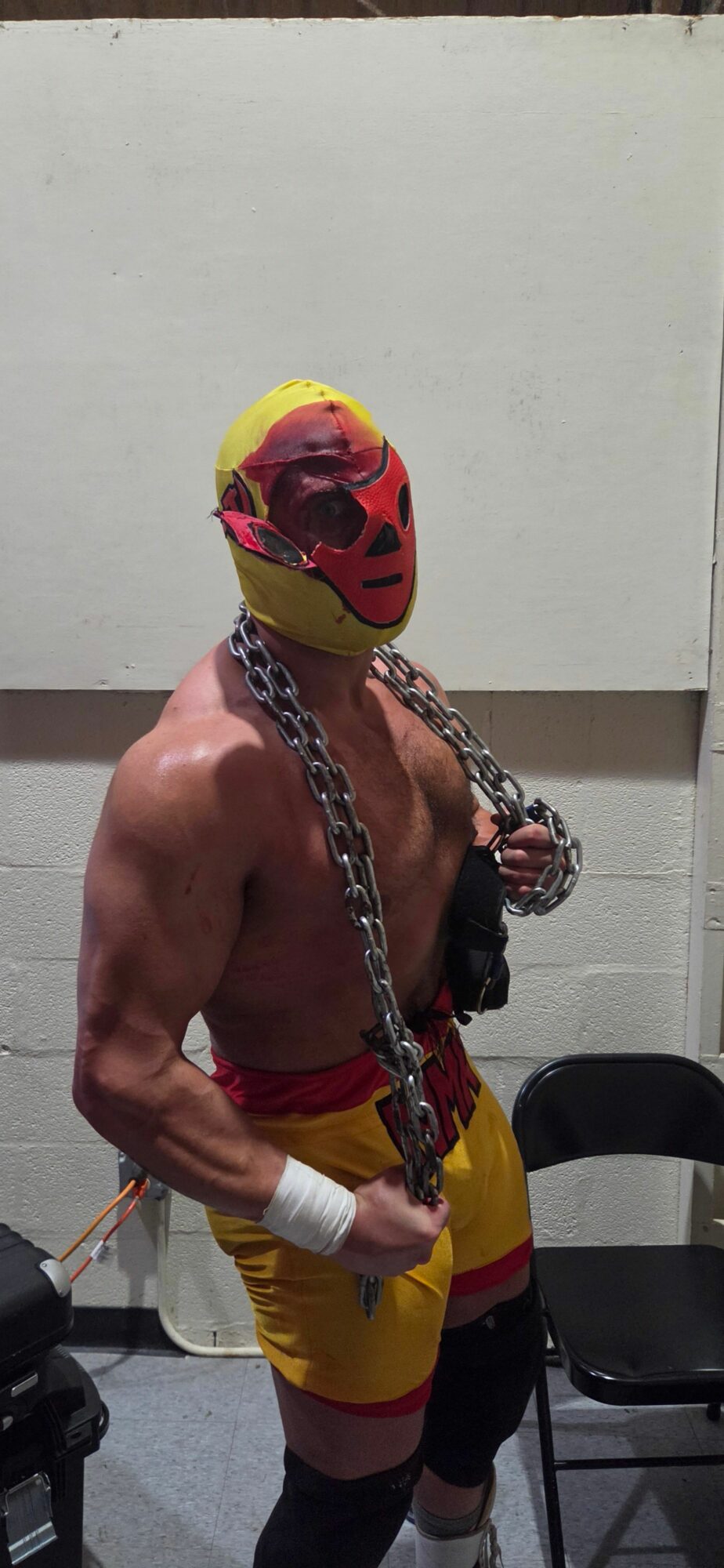 Person wearing a mask and yellow outfit with chains, holding a chain in a room with a white wall.