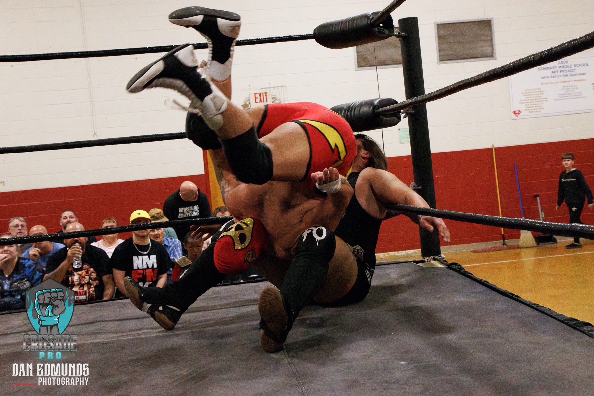 Two wrestlers in a ring, one on top of the other, with spectators watching in the background.