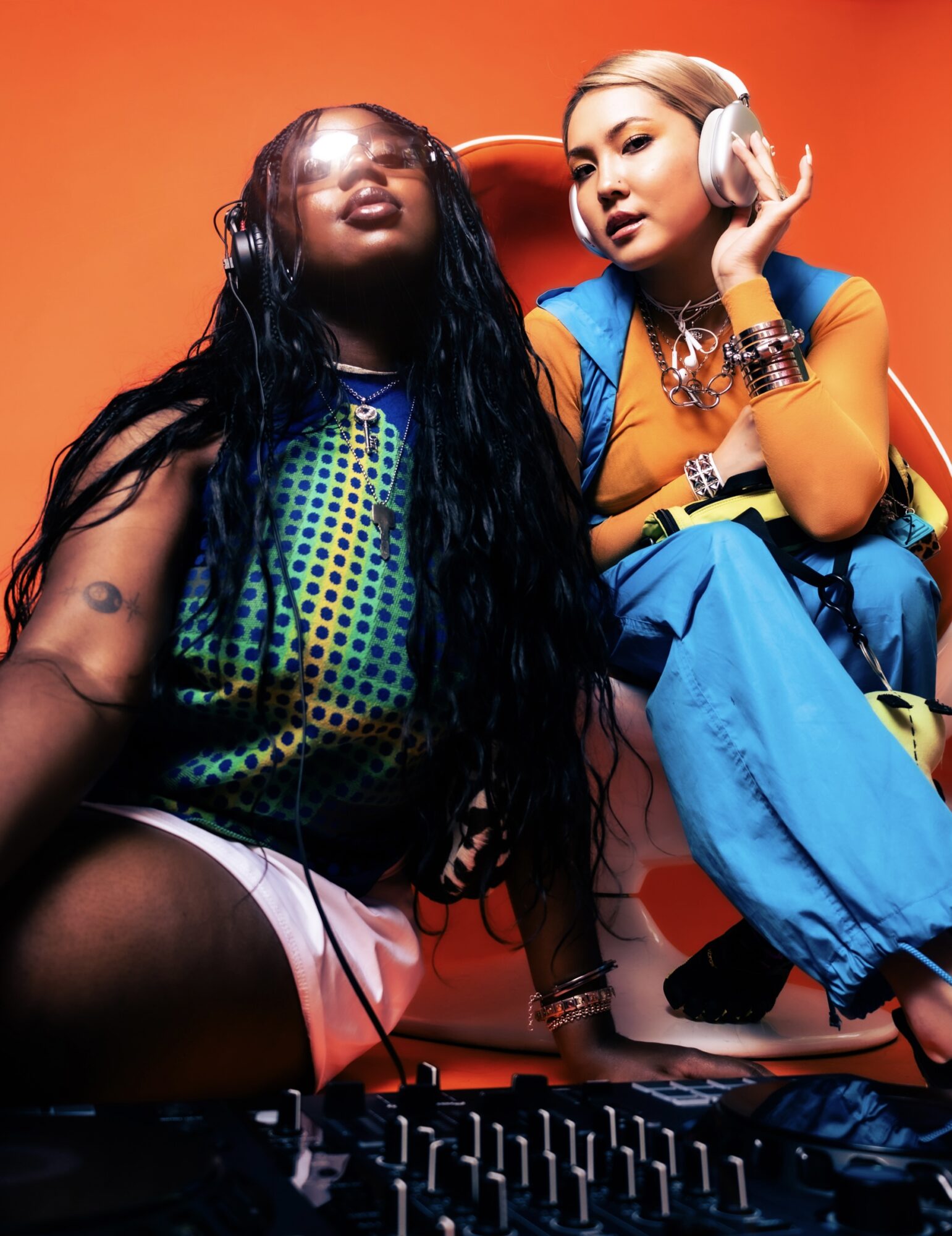 Two women wearing headphones sit against an orange background, one with long dark hair and the other with short blonde hair.