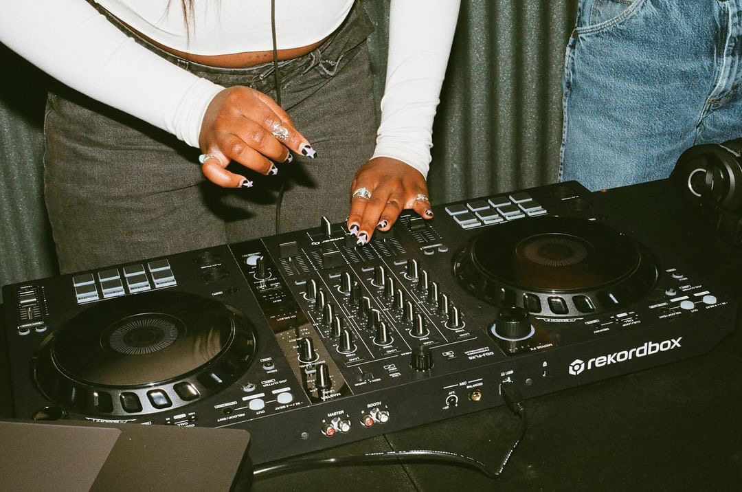 Person's hands on DJ equipment with mixer and turntables, wearing rings and a white long-sleeve shirt.