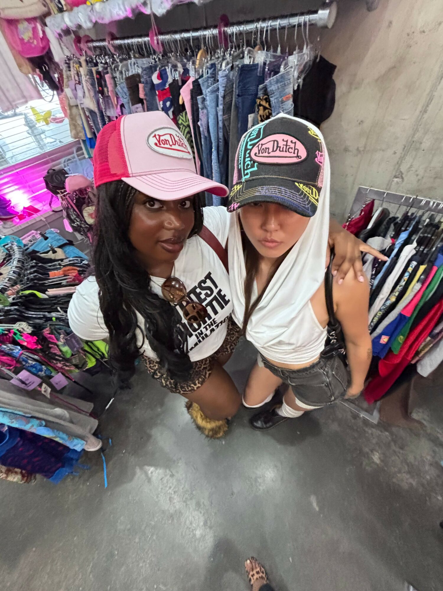Two young women shopping for accessories, one wearing a pink cap and the other a black cap, in a store aisle.