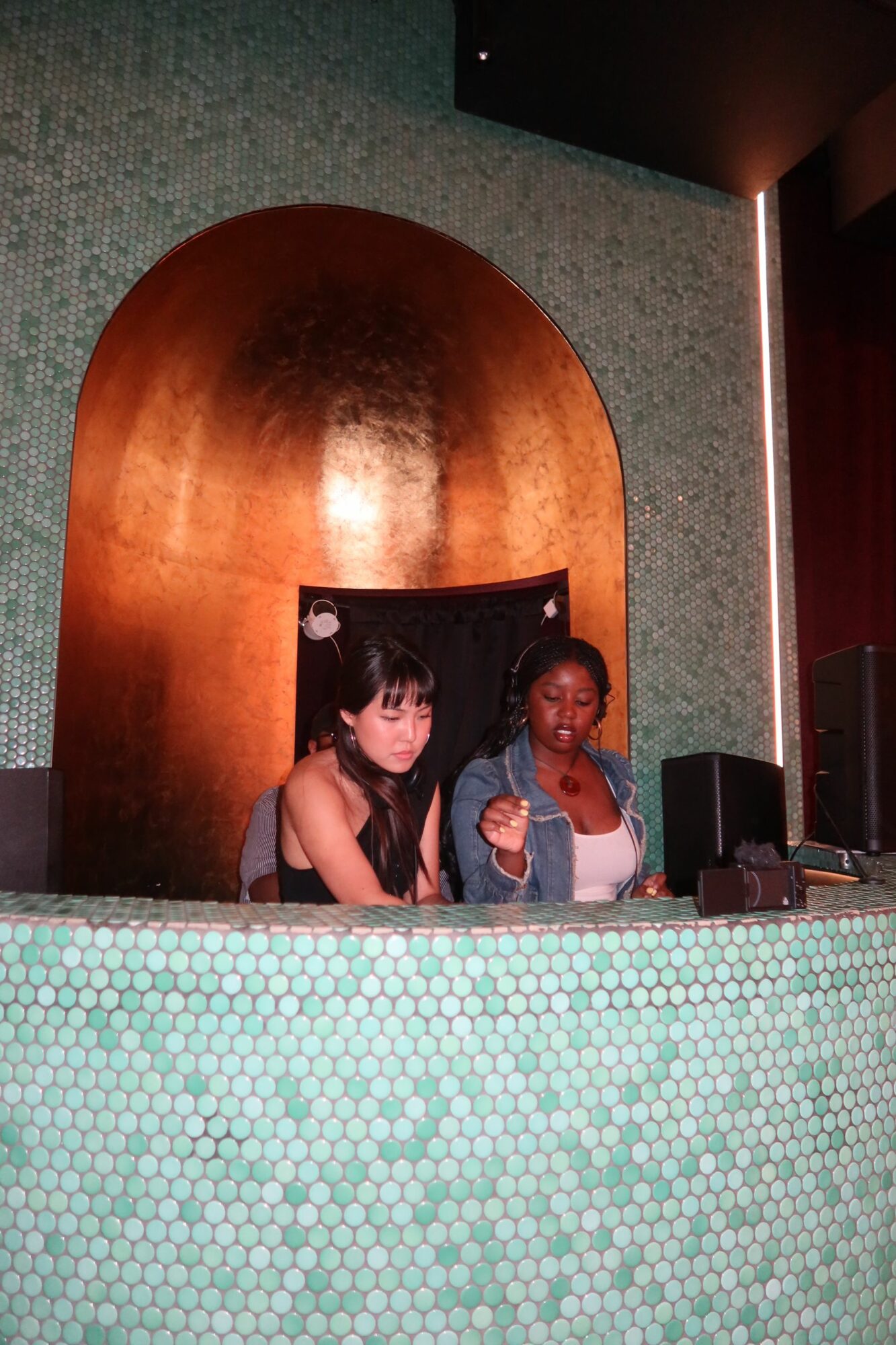 Two women and a man sitting at a bar with a wooden arch behind them, partially visible green tiled surface in front.