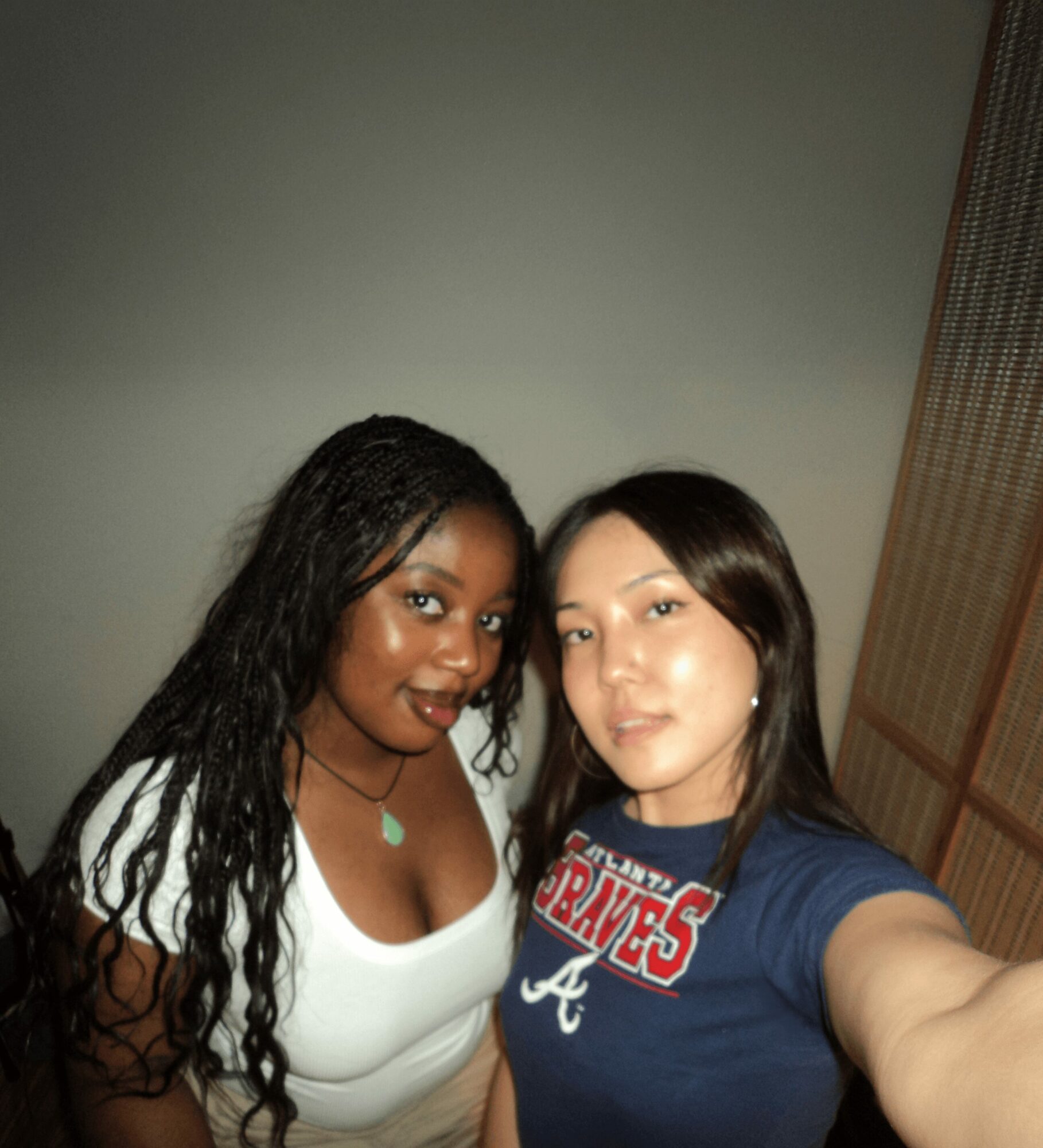Two women taking a selfie indoors, one with long curly hair and the other with straight dark hair, both smiling.