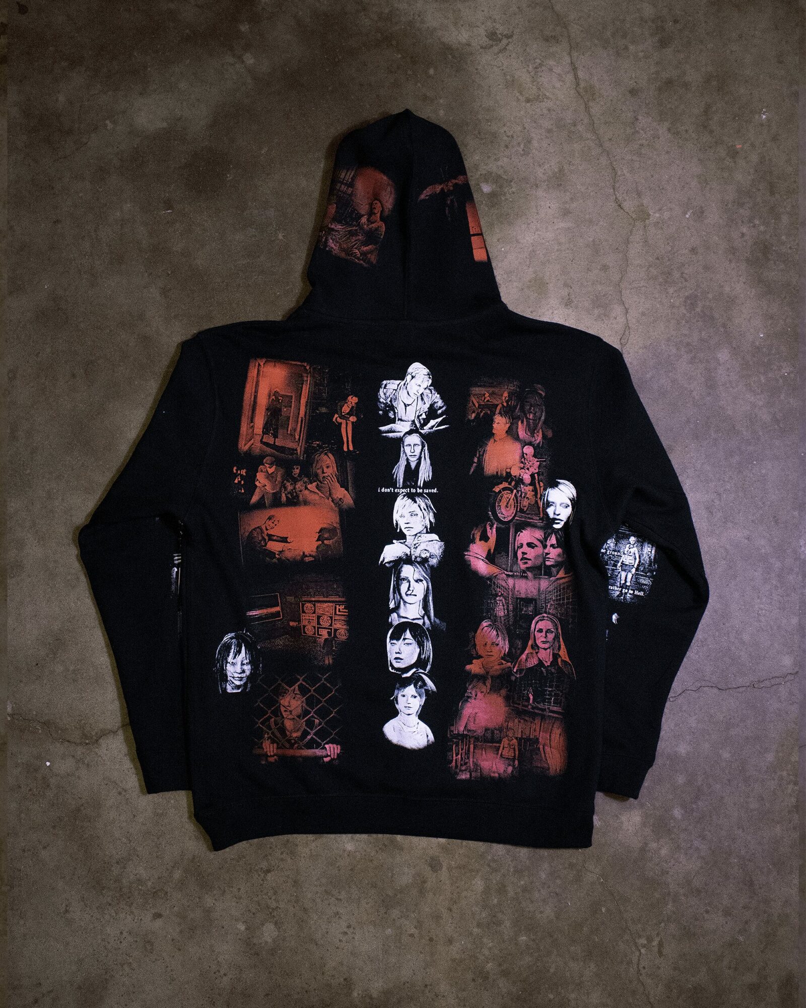Black hoodie with colorful graphic images of faces and scenes, laid flat on a concrete surface.