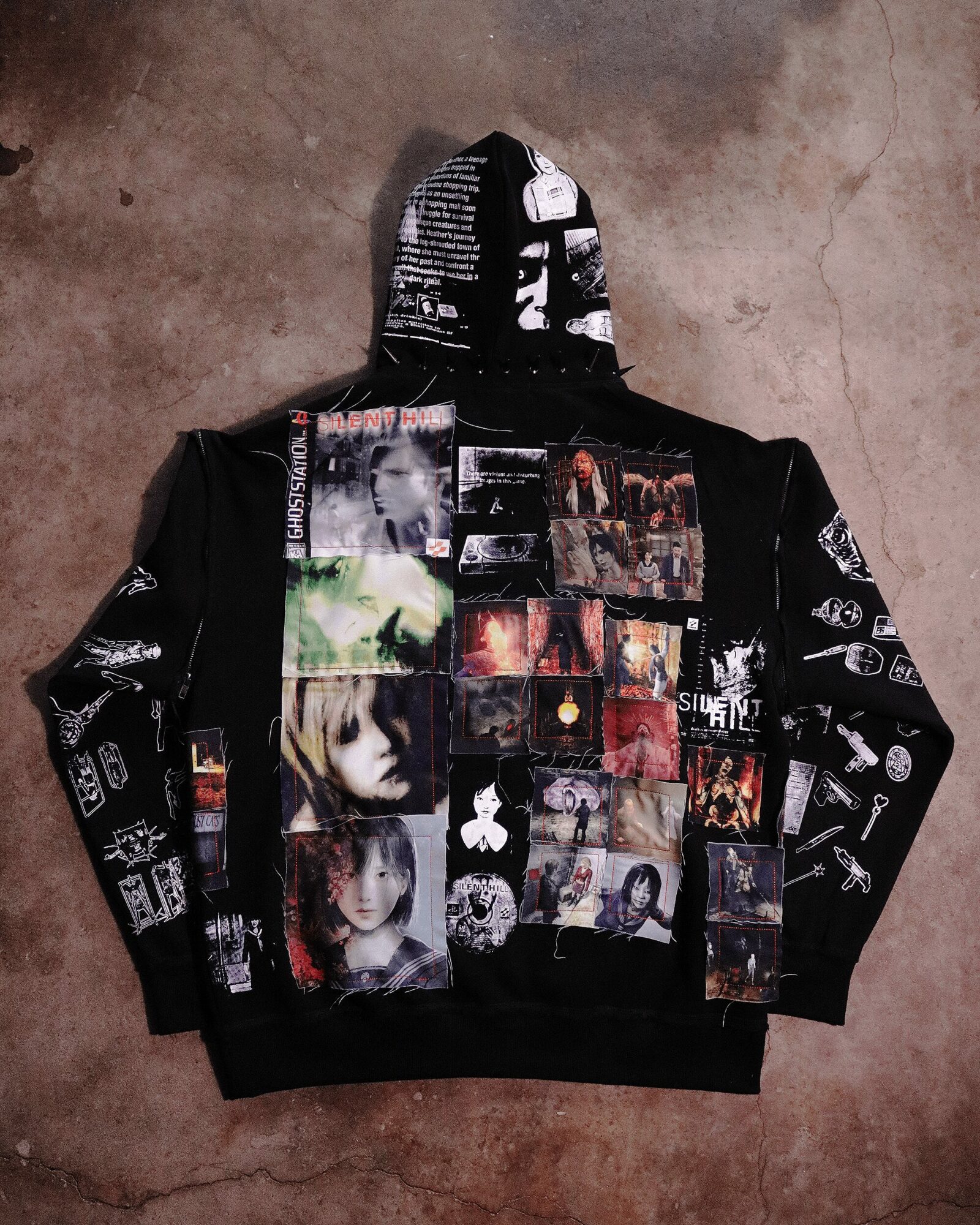 Black hoodie with collage of images and text, laid flat on a concrete surface, with the hood up.