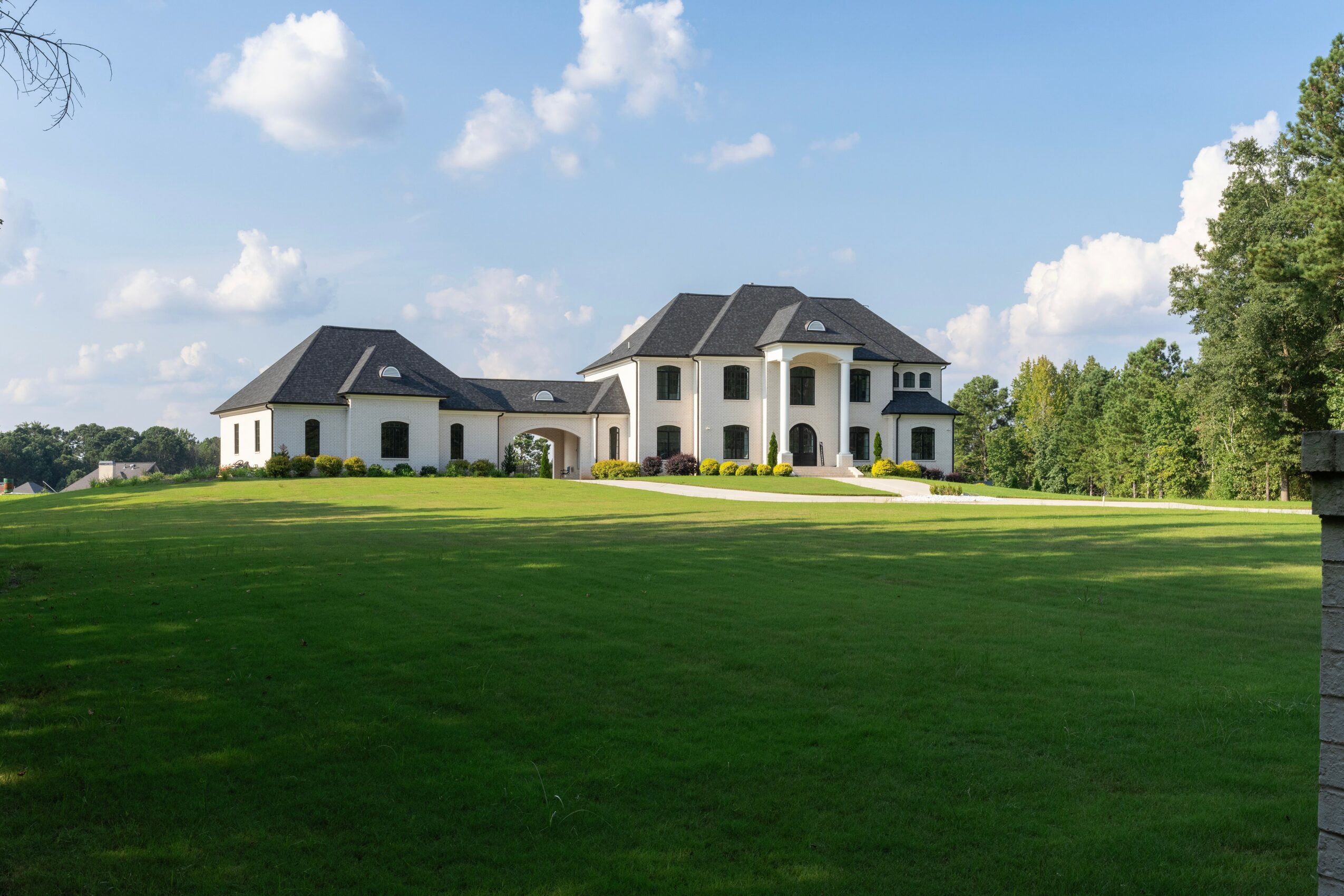 Large white house with multiple sections and a dark roof, surrounded by a green lawn and trees, under a partly cloudy sky.