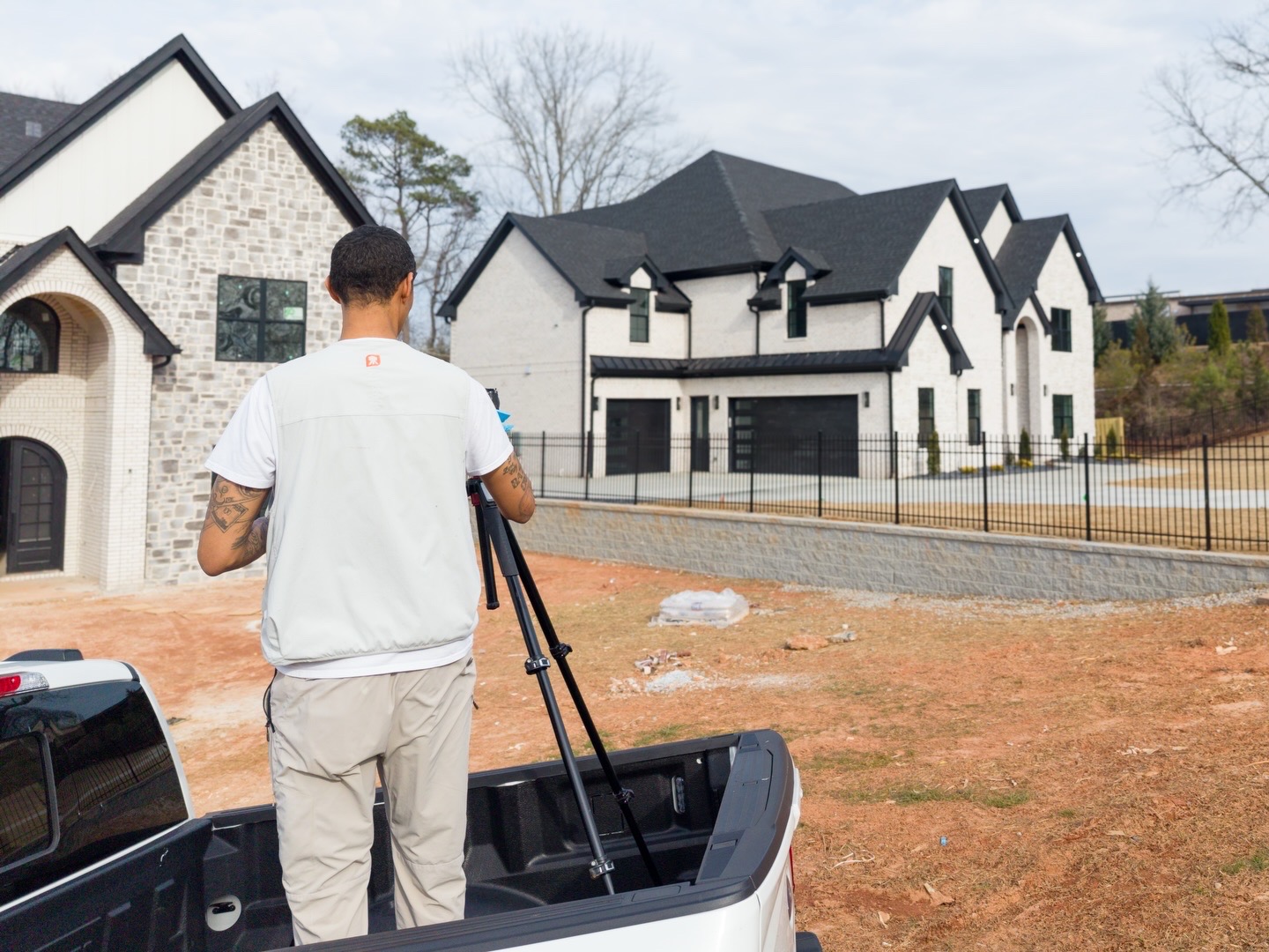 Man with camera equipment stands in the back of a truck, overlooking modern white houses with black roofs and a fenced yard.