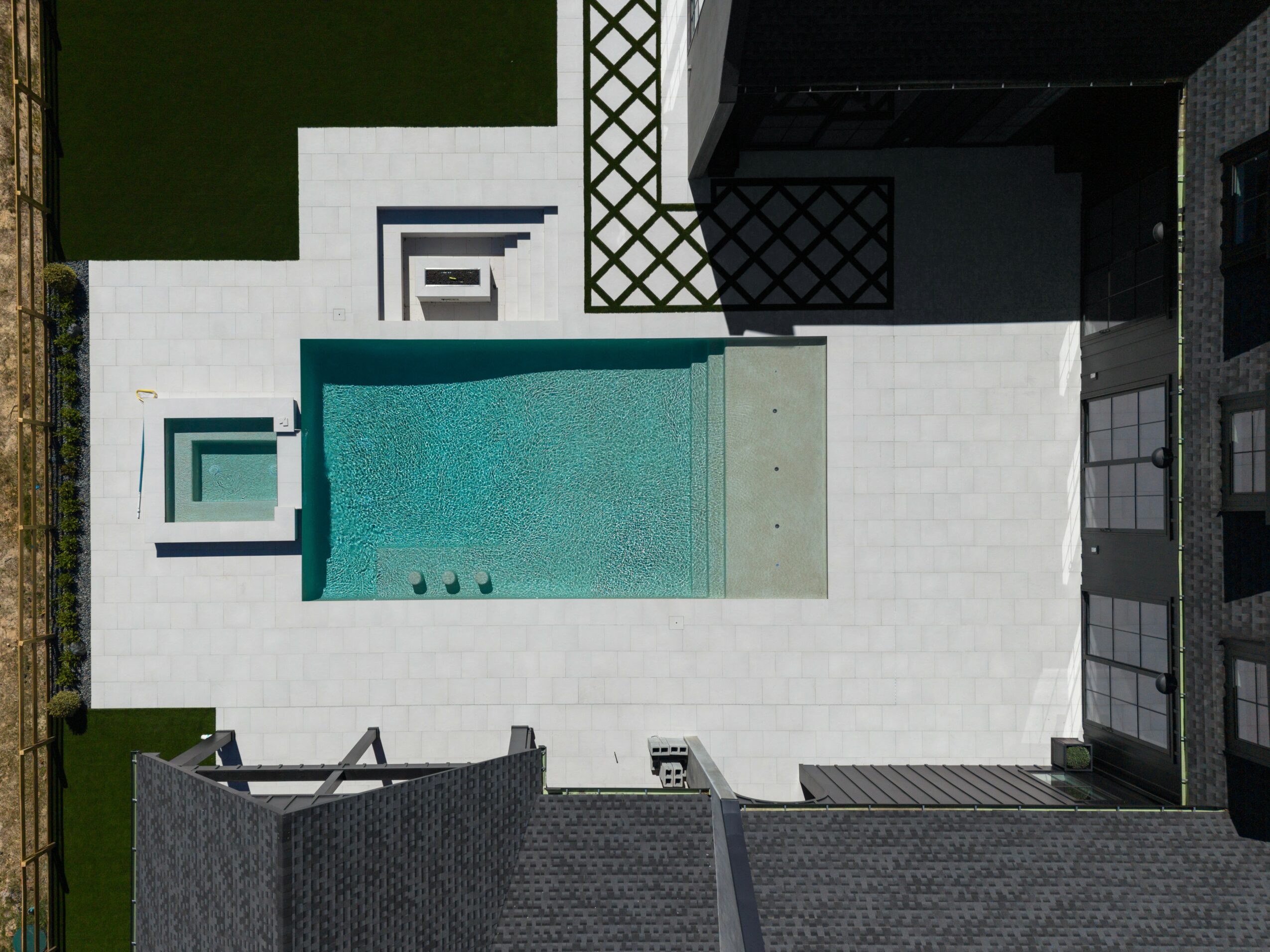 Aerial view of a modern backyard with a rectangular swimming pool, white tiled patio, and surrounding buildings.