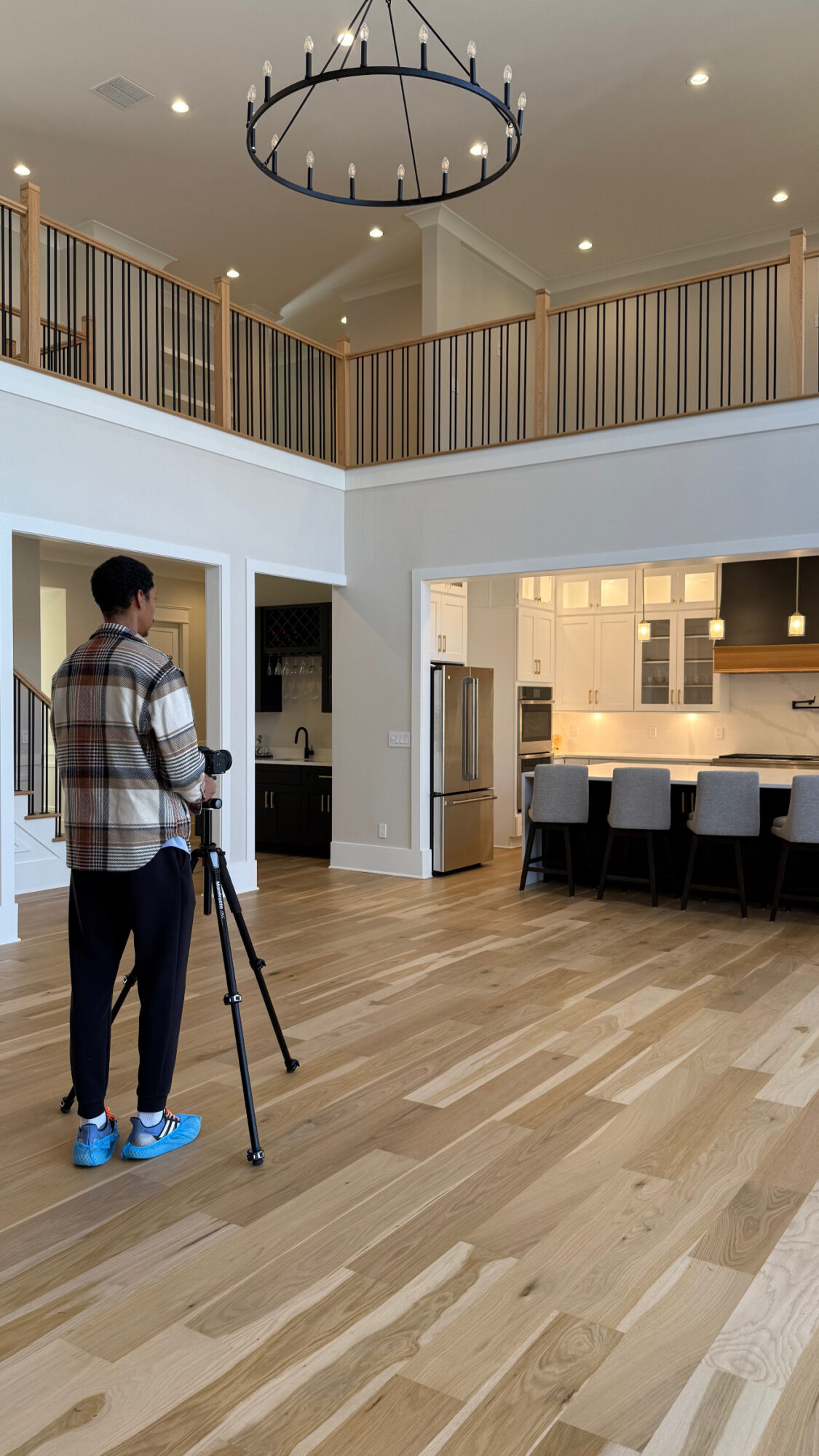 Person with camera tripod in spacious open-concept living area with kitchen and dining space, hardwood floors, and high ceiling.
