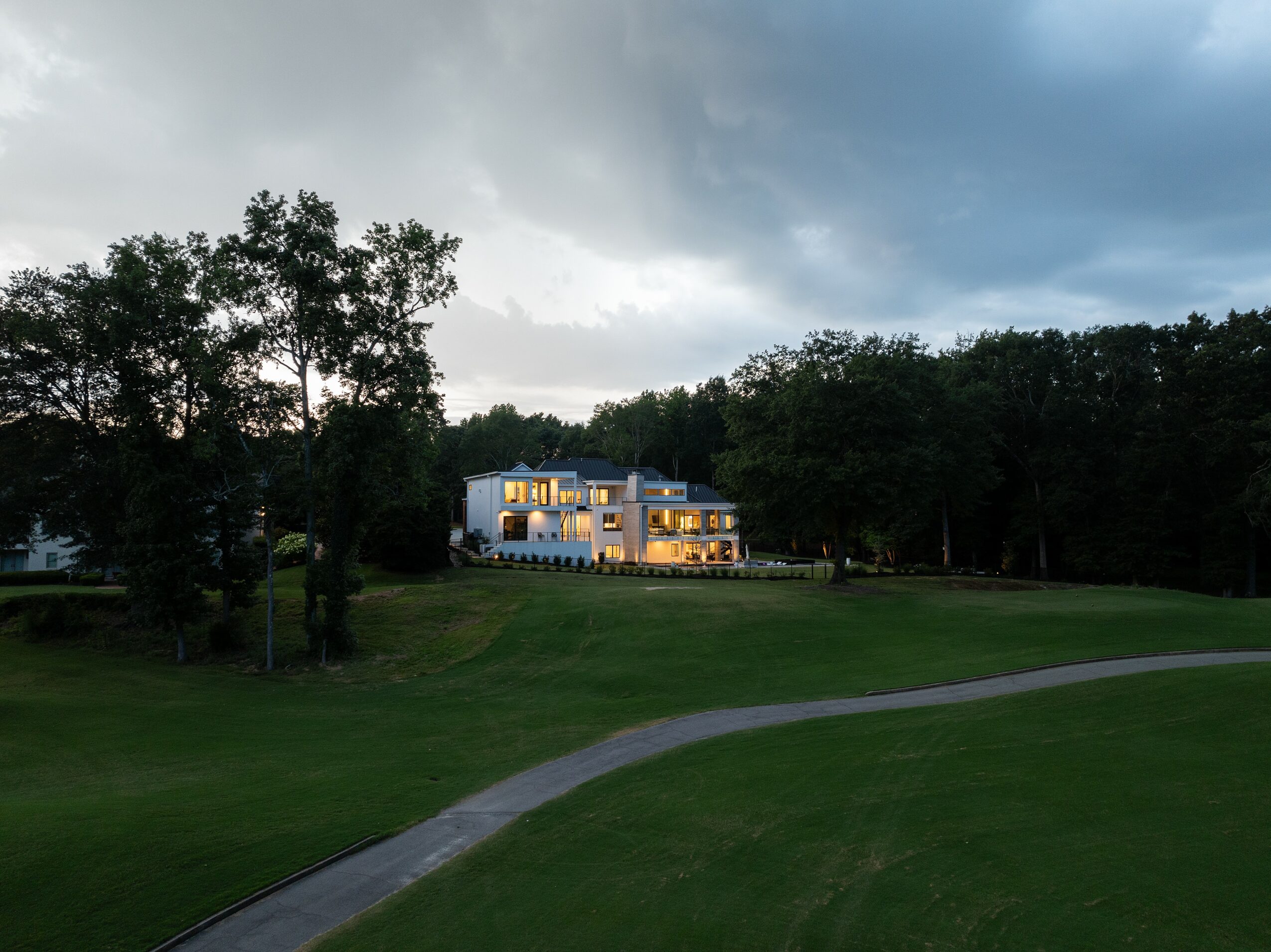 Large white house with multiple windows illuminated, surrounded by trees and a grassy area, under a cloudy sky.