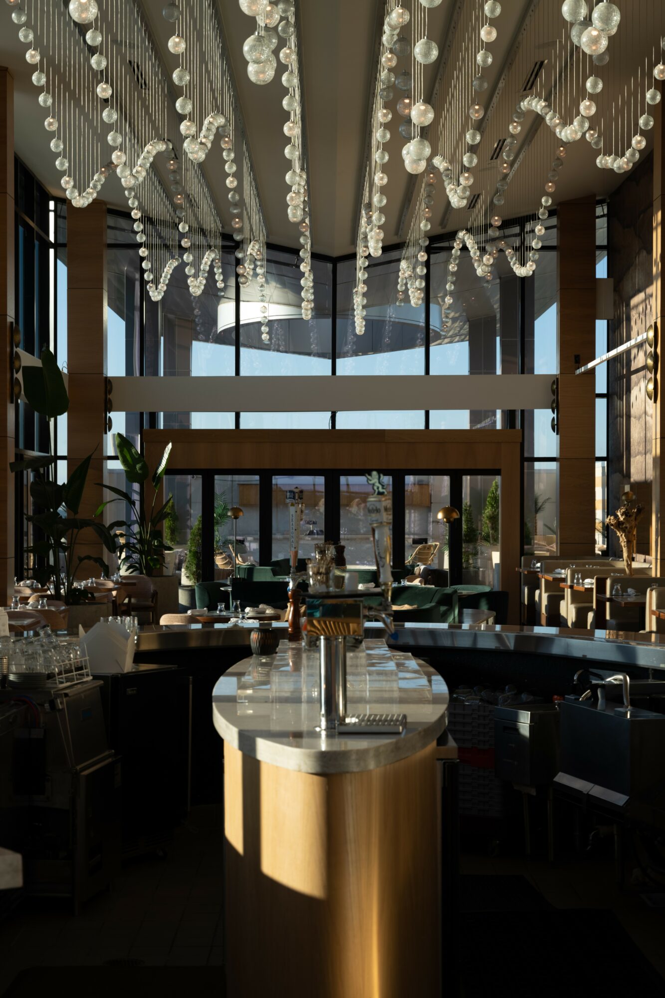 Interior of a restaurant with hanging light fixtures, large windows, and a central table with a bottle and glass, sunlight streaming in.