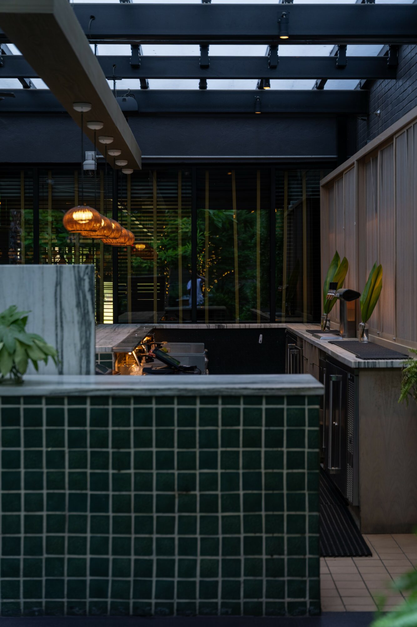 Indoor space with a tiled water feature, hanging lights, and plants, viewed through glass walls with a dark ceiling.