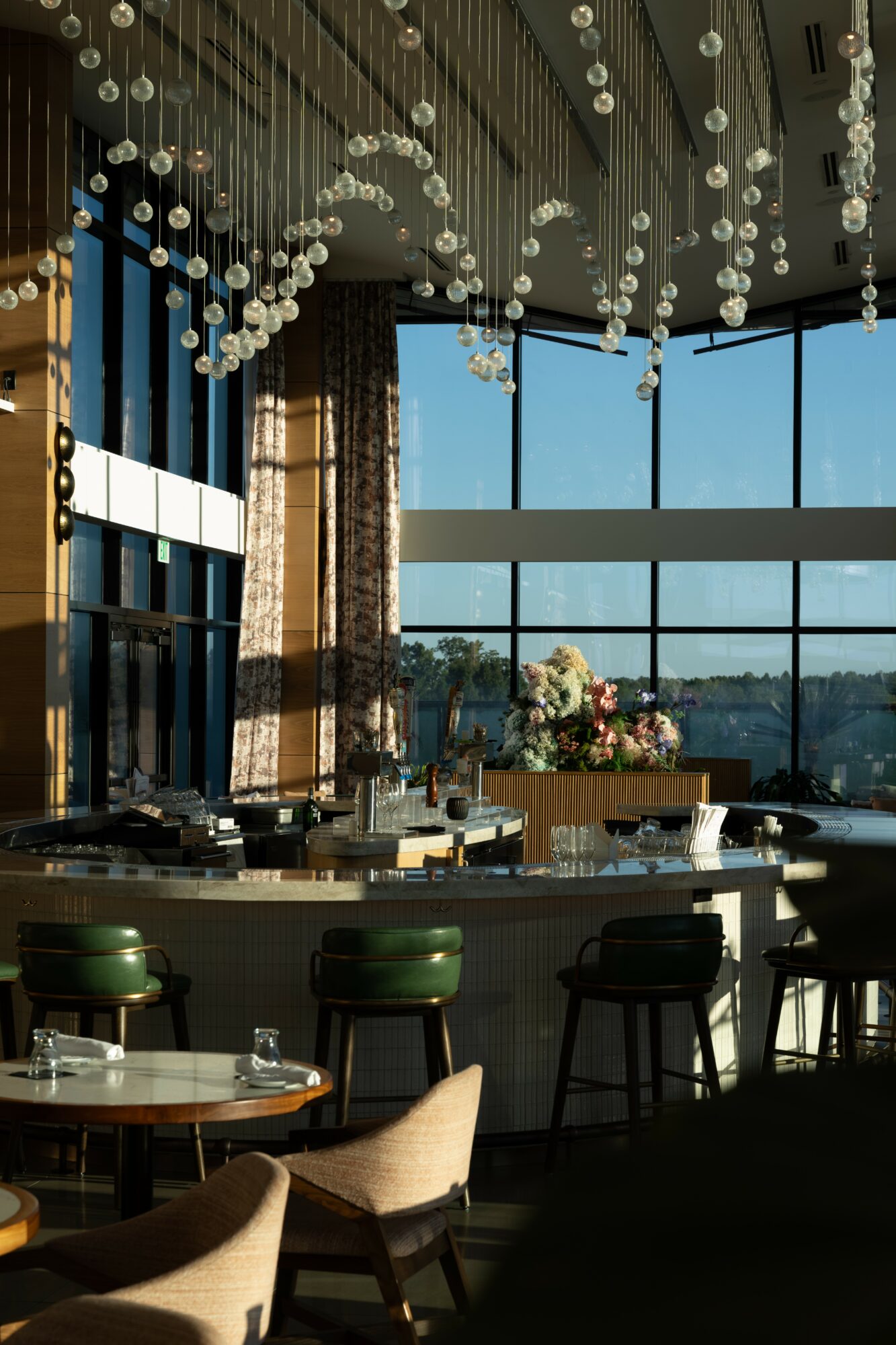Interior of a restaurant with hanging lights, large windows, tables, and chairs, with a city view outside.