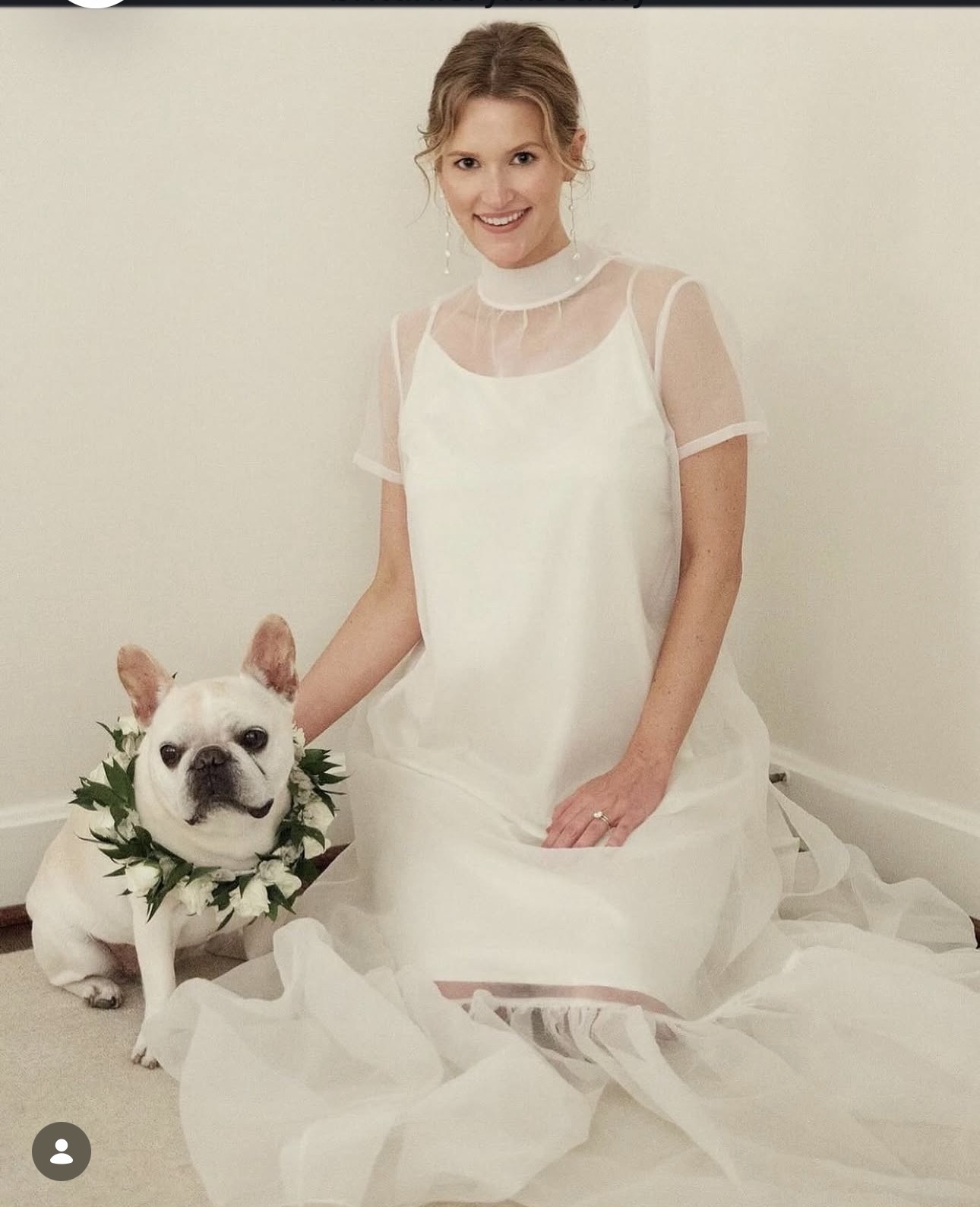 Woman in white dress sitting on floor with a dog wearing a floral wreath, smiling at camera.