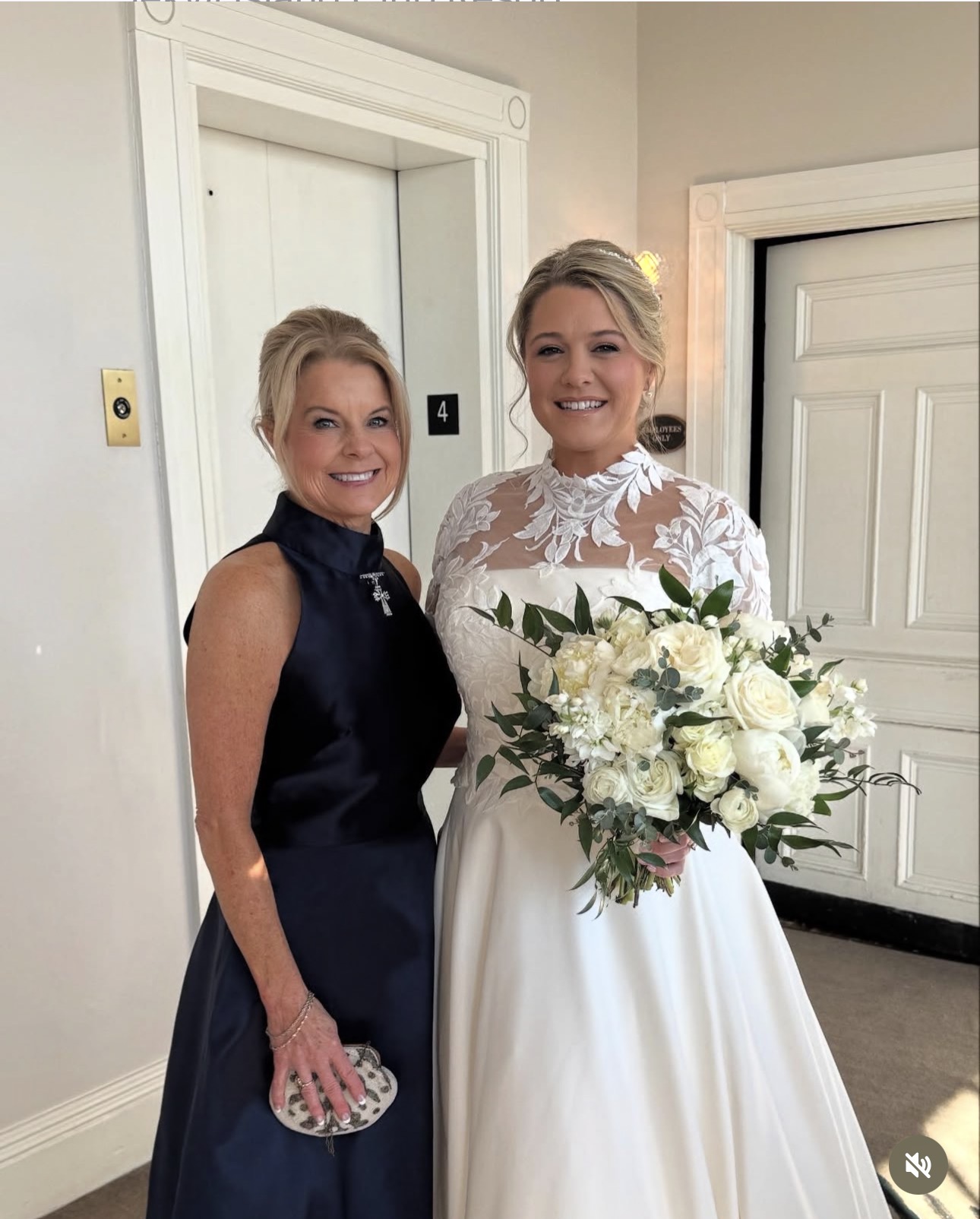 Two women, one in a dark dress and the other in a white wedding gown, smiling indoors with a bouquet of white flowers.