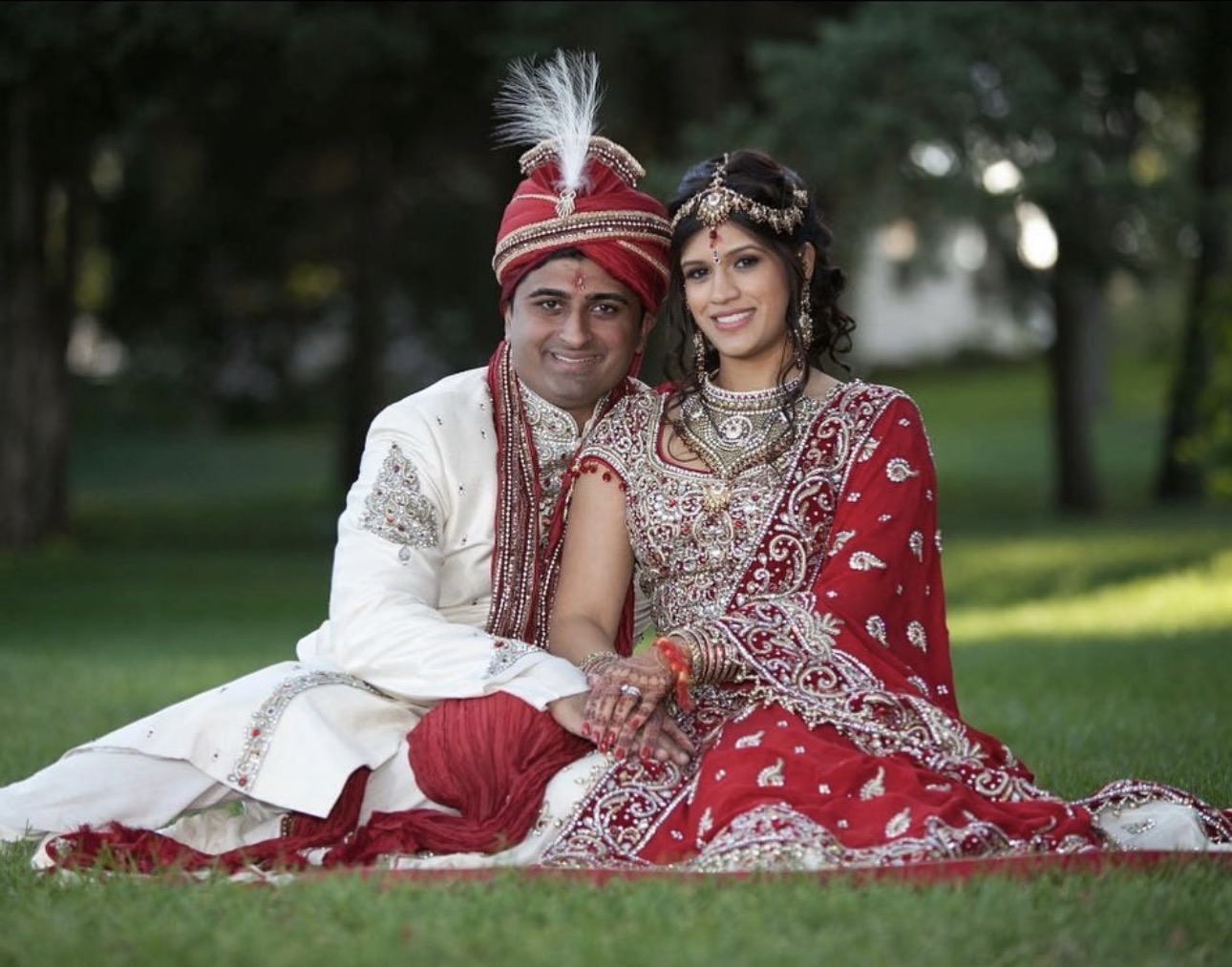 A couple in traditional wedding attire sitting on grass outdoors, smiling, with trees in the background.