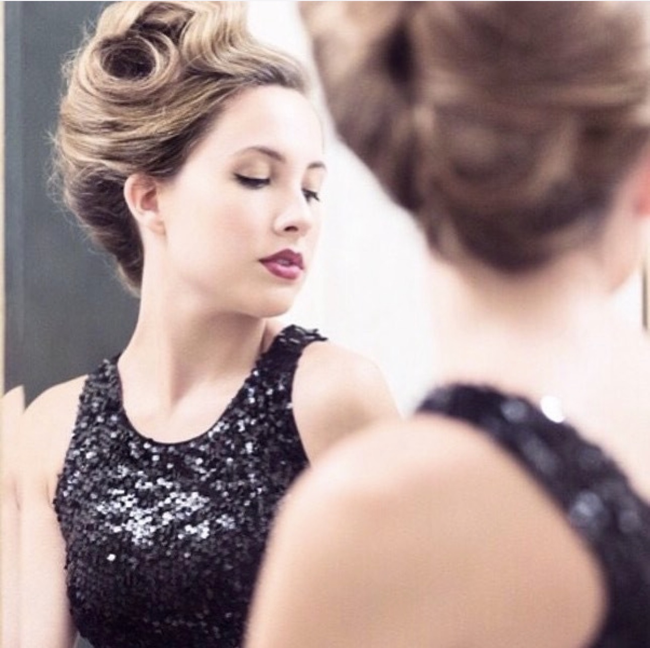 Woman with styled hair and makeup looking at her reflection in a mirror, wearing a black sequined dress.