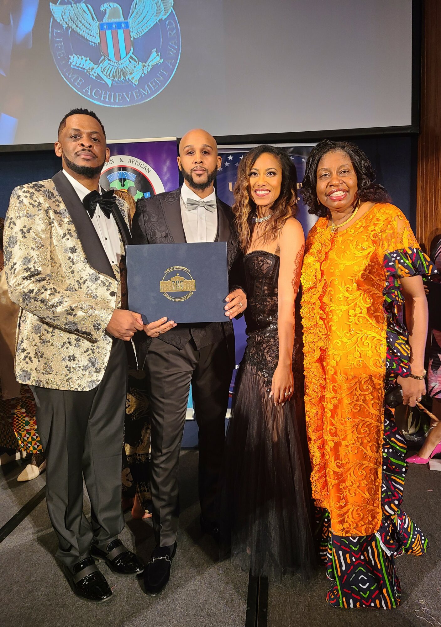 Four people dressed in formal and colorful attire standing together indoors, with a large screen displaying a logo behind them.