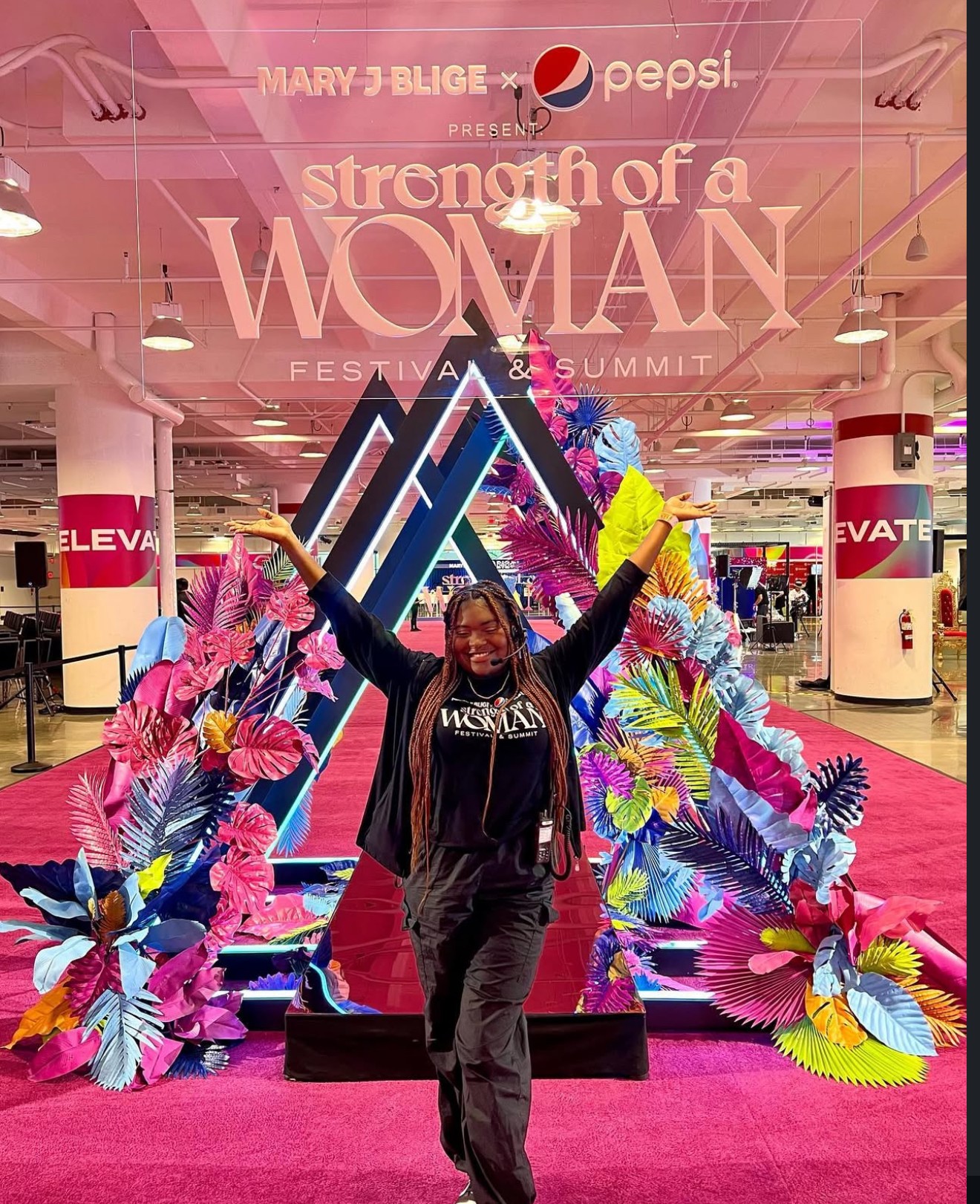 Person standing in front of a colorful floral display at an event, arms raised, with a pink carpet and event signage in the background.