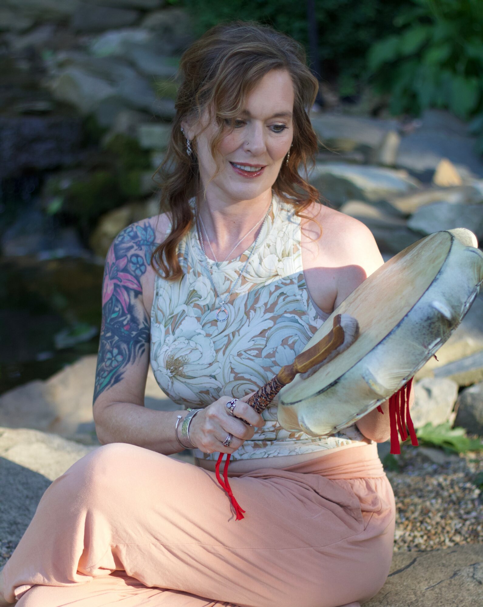 Woman with wavy hair, wearing a sleeveless top, sitting outdoors near rocks, holding a tambourine, smiling.
