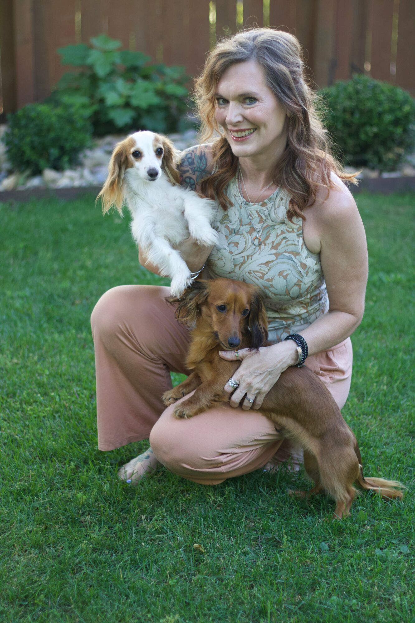 Woman sitting on grass with two dogs, smiling, in a backyard with a wooden fence and green plants.