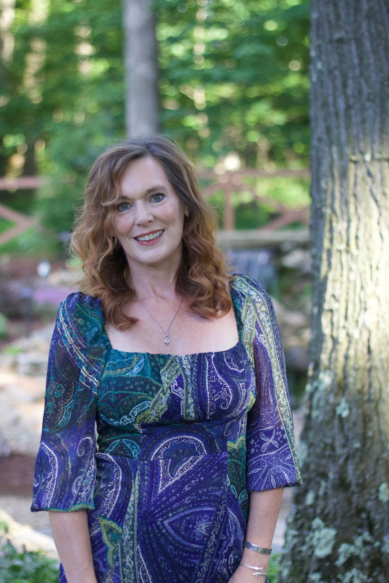 Woman with wavy hair smiling outdoors in a forest setting, wearing a colorful patterned dress with trees in the background.