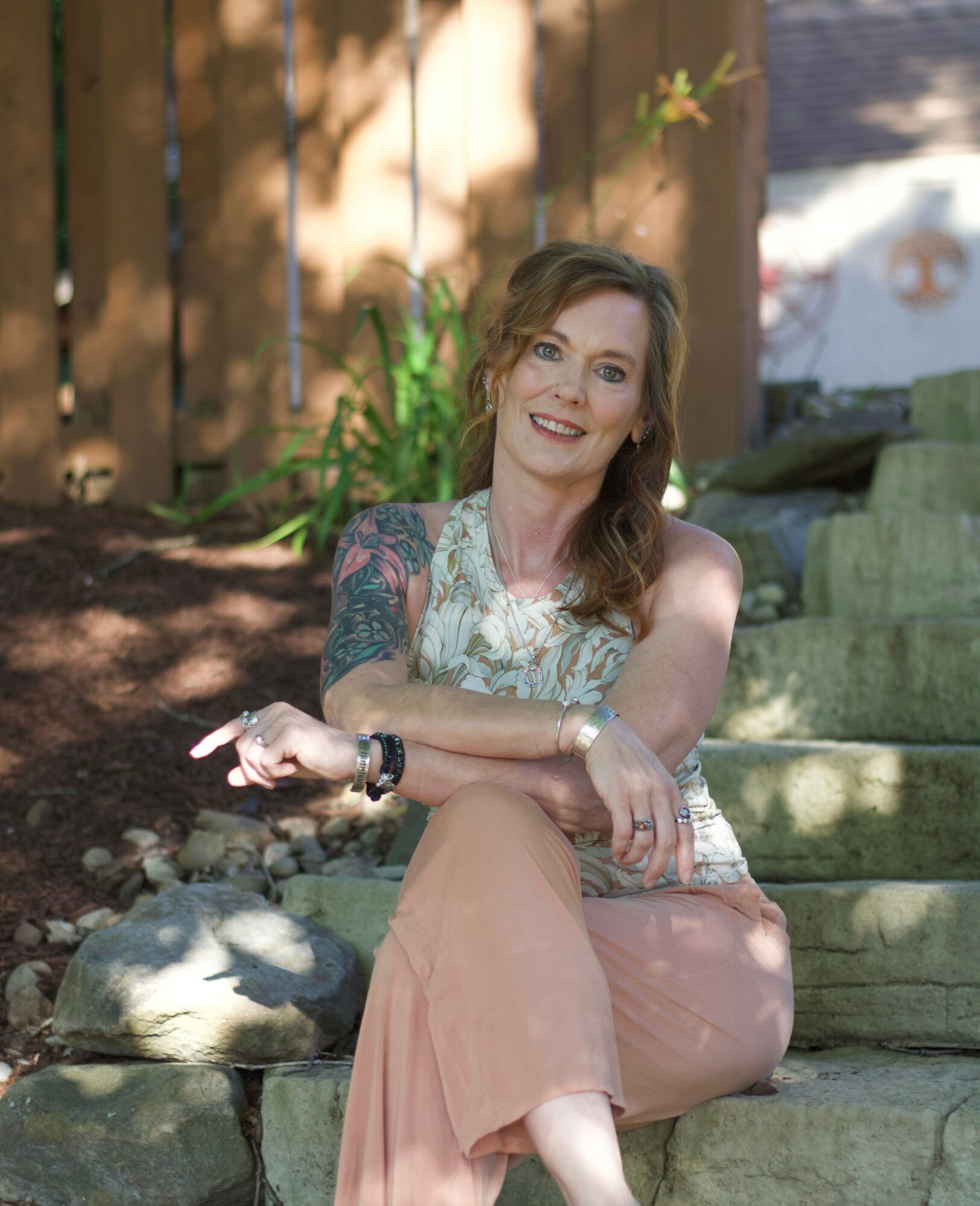Woman with wavy hair sitting on outdoor stone steps, smiling, wearing a sleeveless top and peach-colored pants, with a wooden fence and plants in background.