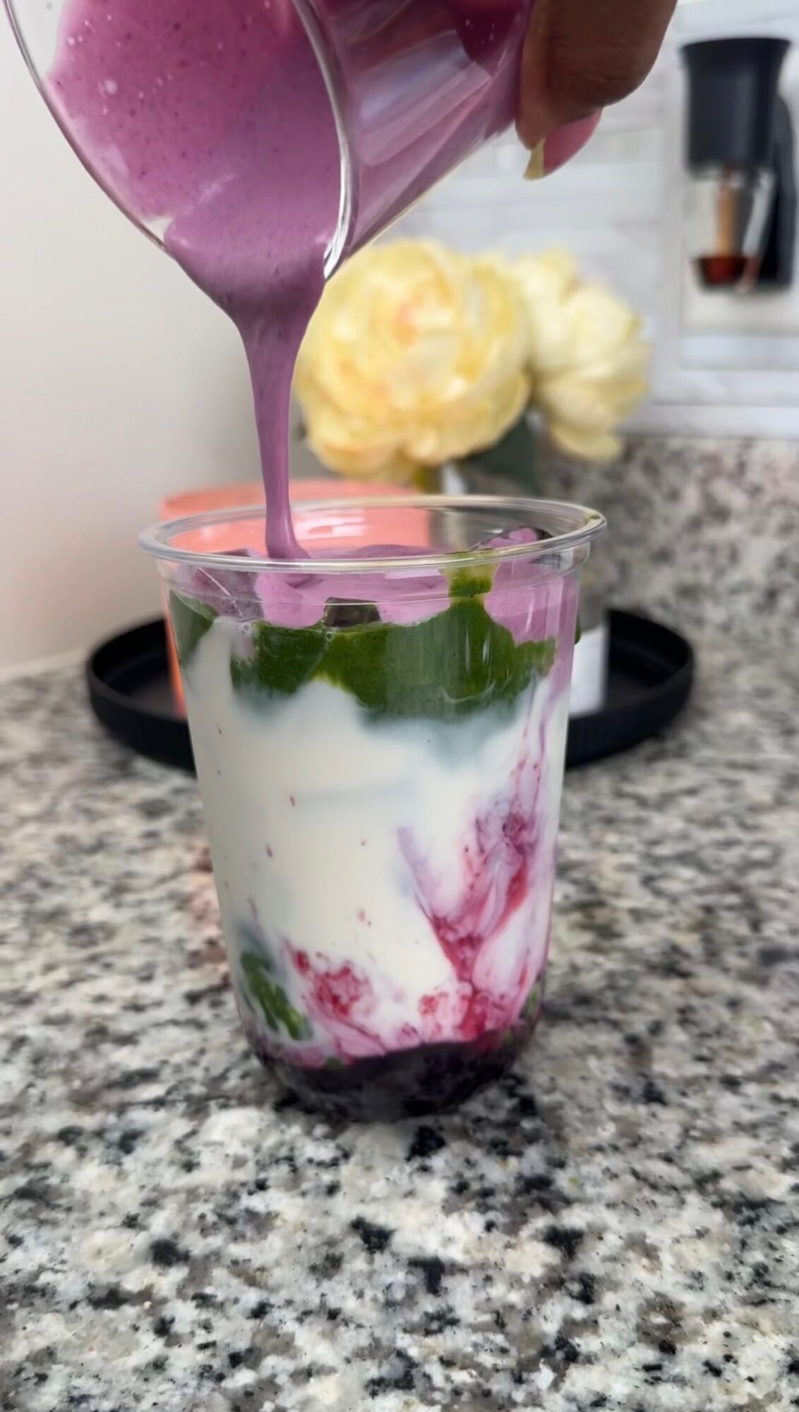 Pouring purple smoothie into a glass with layered yogurt and fruit, on a kitchen countertop.
