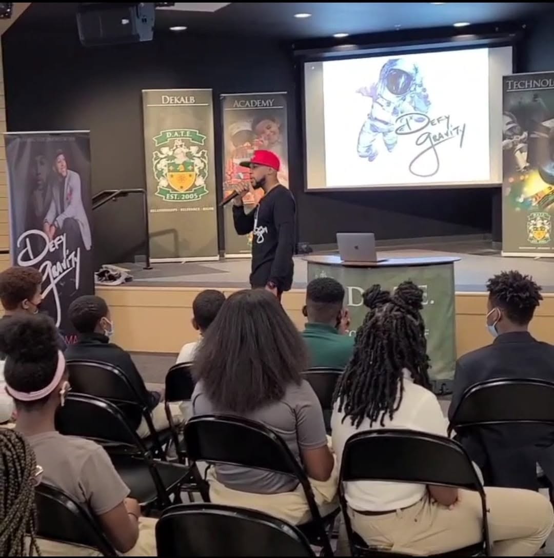 Person in red cap speaking to seated audience in a classroom or seminar room with banners and a screen behind.