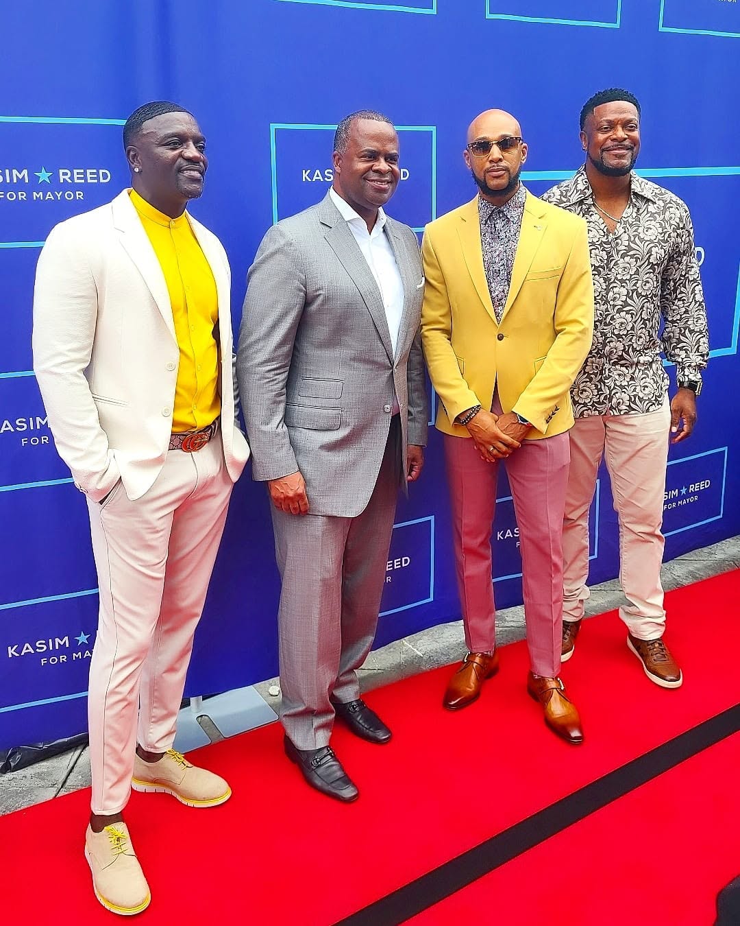 Four men in formal and semi-formal attire standing on a red carpet in front of a blue backdrop with text.
