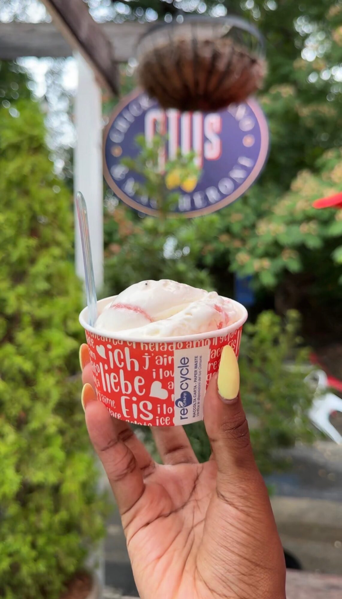 Person holding a cup of ice cream outdoors with a sign and trees in the background.