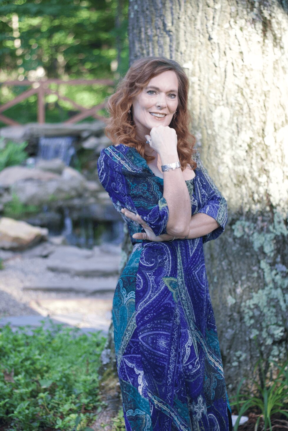 Woman with curly hair smiling outdoors, standing near a tree with a pond in the background.