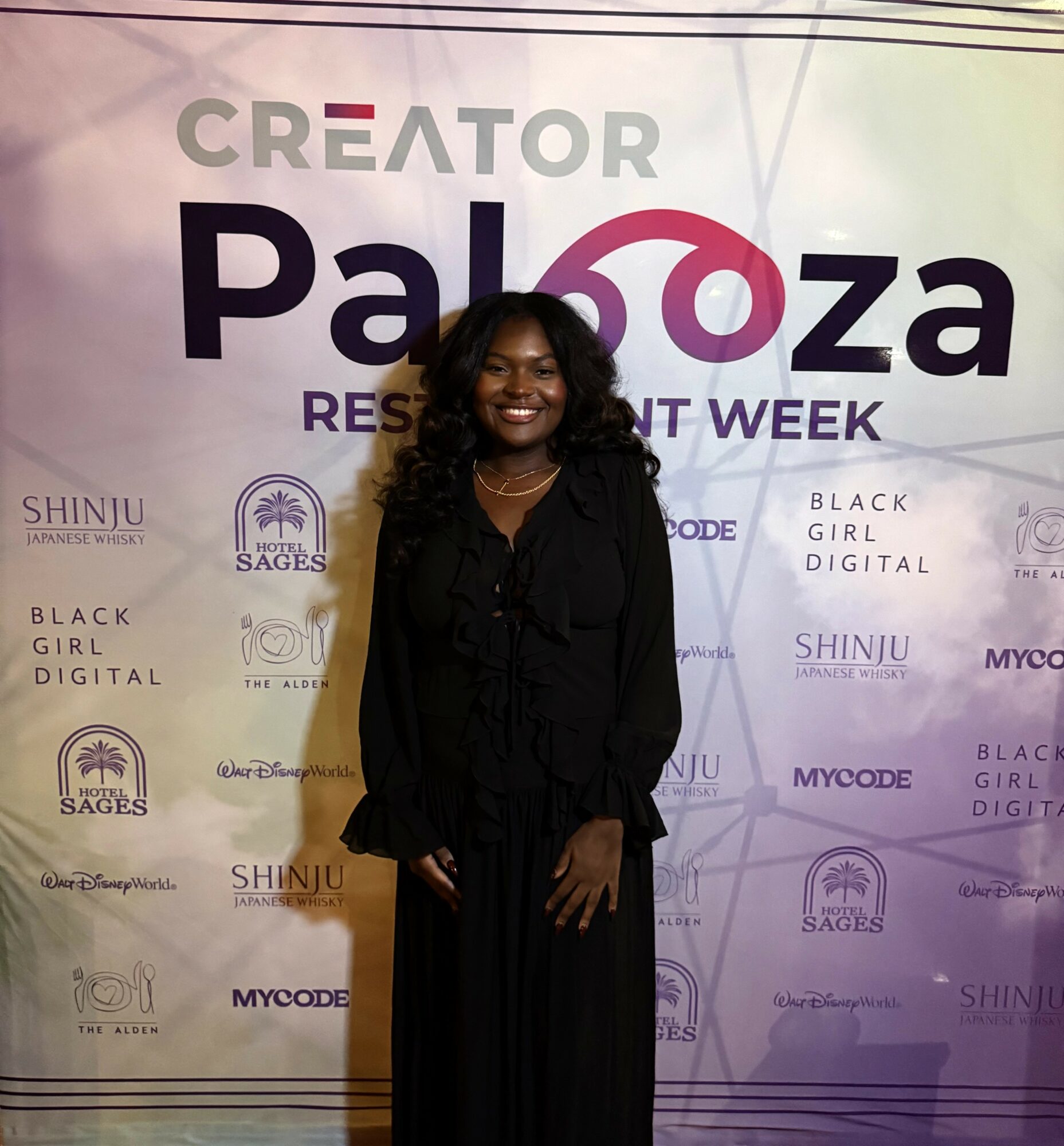 Smiling woman in black standing in front of a purple event backdrop with text and logos.