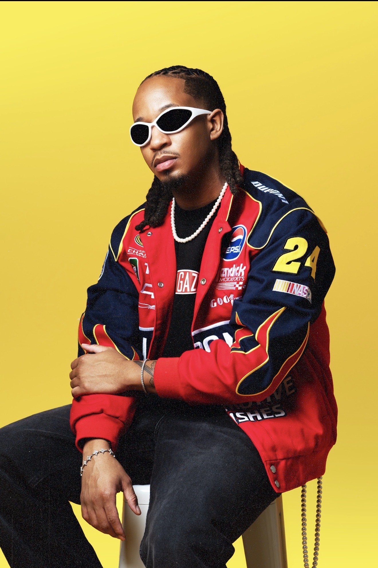 Person wearing sunglasses and a racing jacket, sitting against a yellow background.