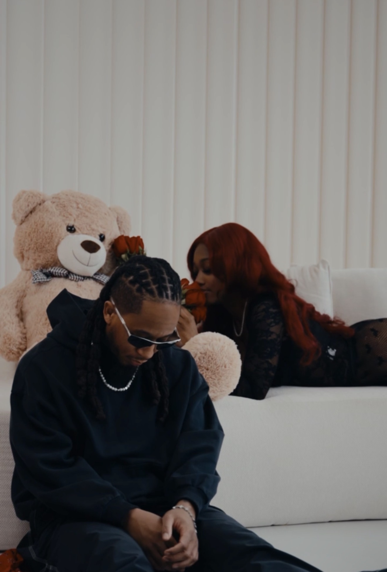 Two people sitting on a white couch with a large teddy bear, one with braided hair and sunglasses, the other with long red hair.
