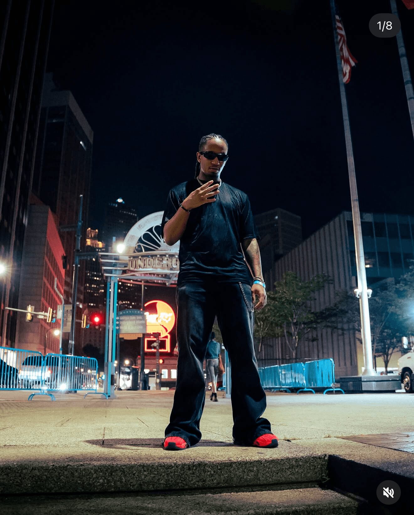 Person standing on city street at night, wearing dark clothing and sunglasses, with buildings and a Ferris wheel in background.