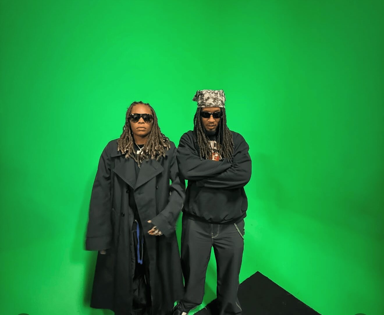 Two people with dreadlocks and sunglasses stand against a green background, one with arms crossed, the other with hands in pockets.