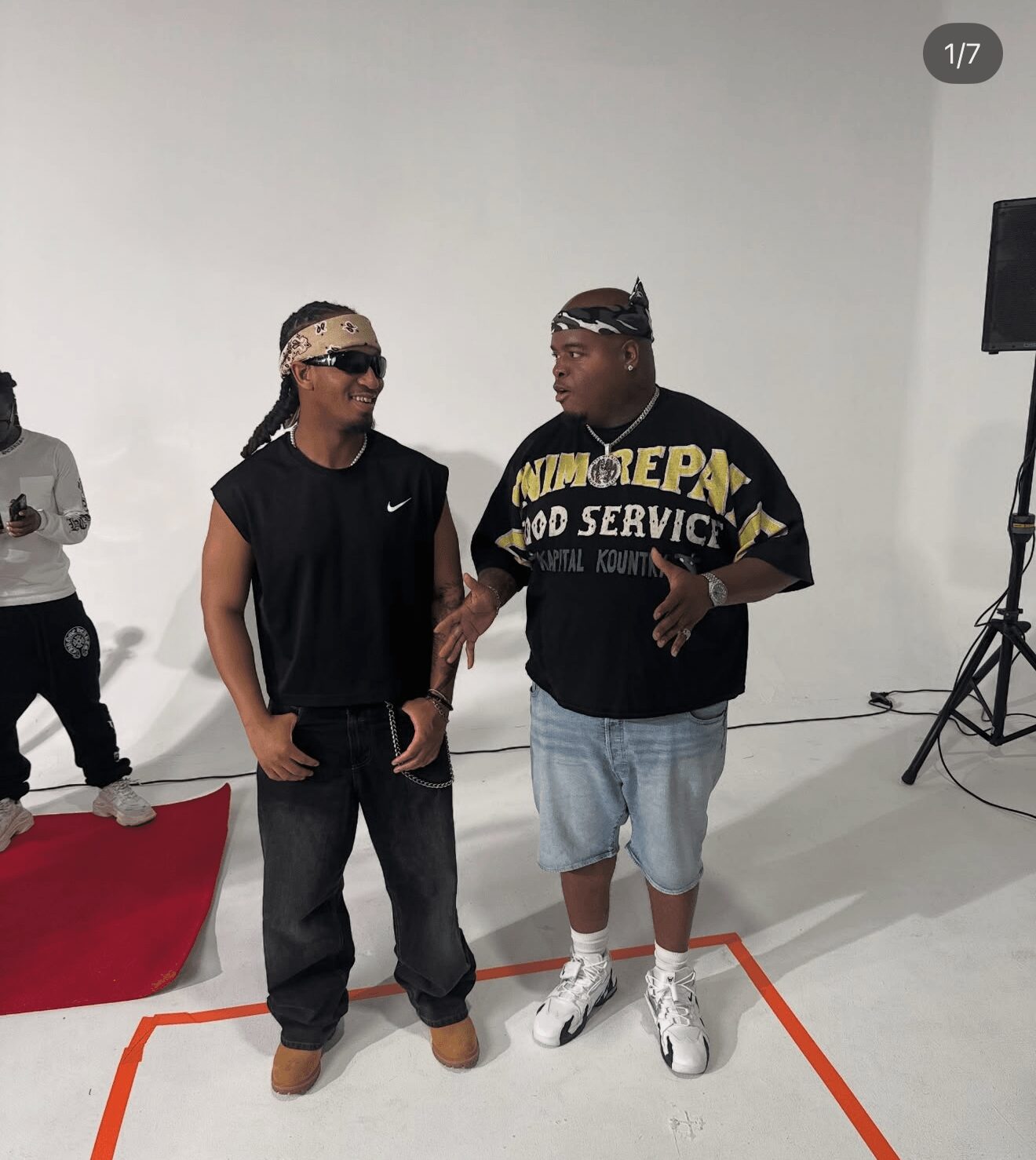 Two men stand and talk in a white studio with a red platform and a speaker on a stand. One wears sunglasses and a bandana, the other a T-shirt and shorts.