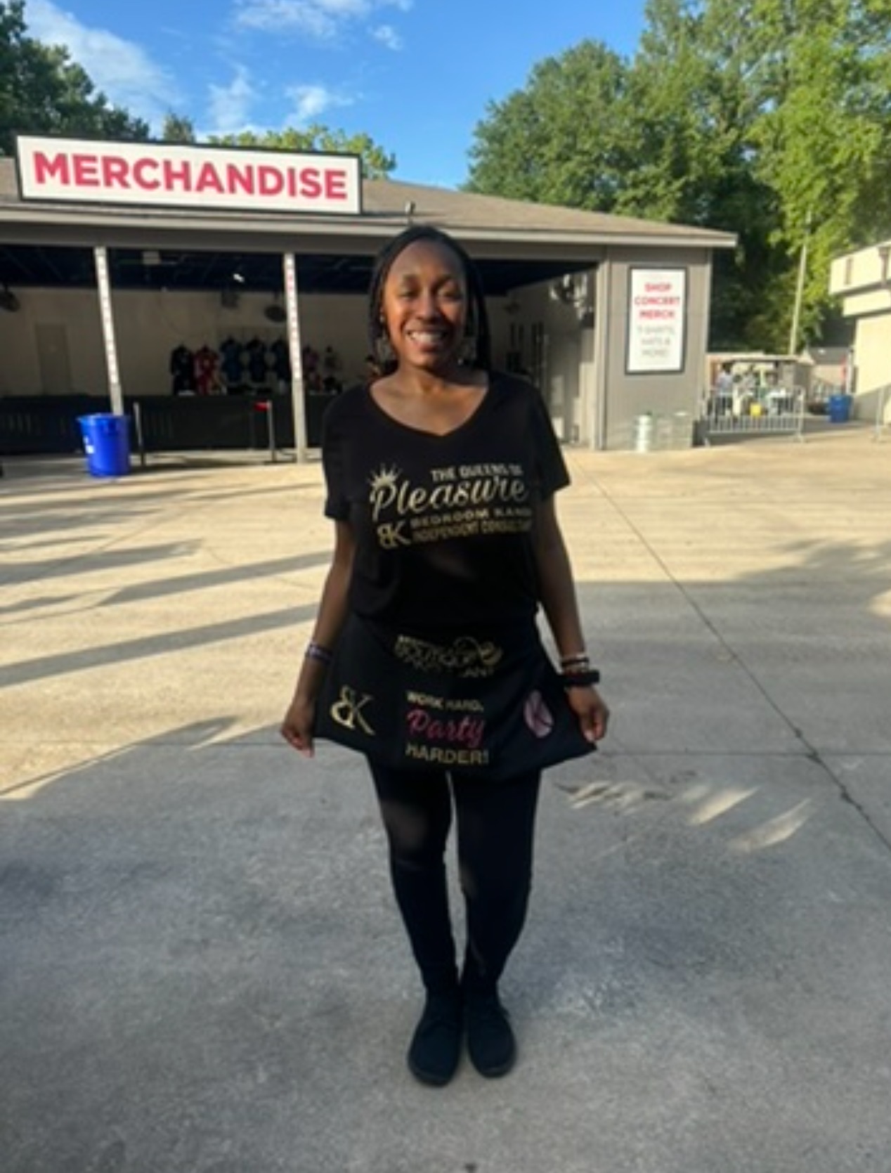 Smiling woman standing outdoors in front of a merchandise stand, wearing black clothing and holding a bag.