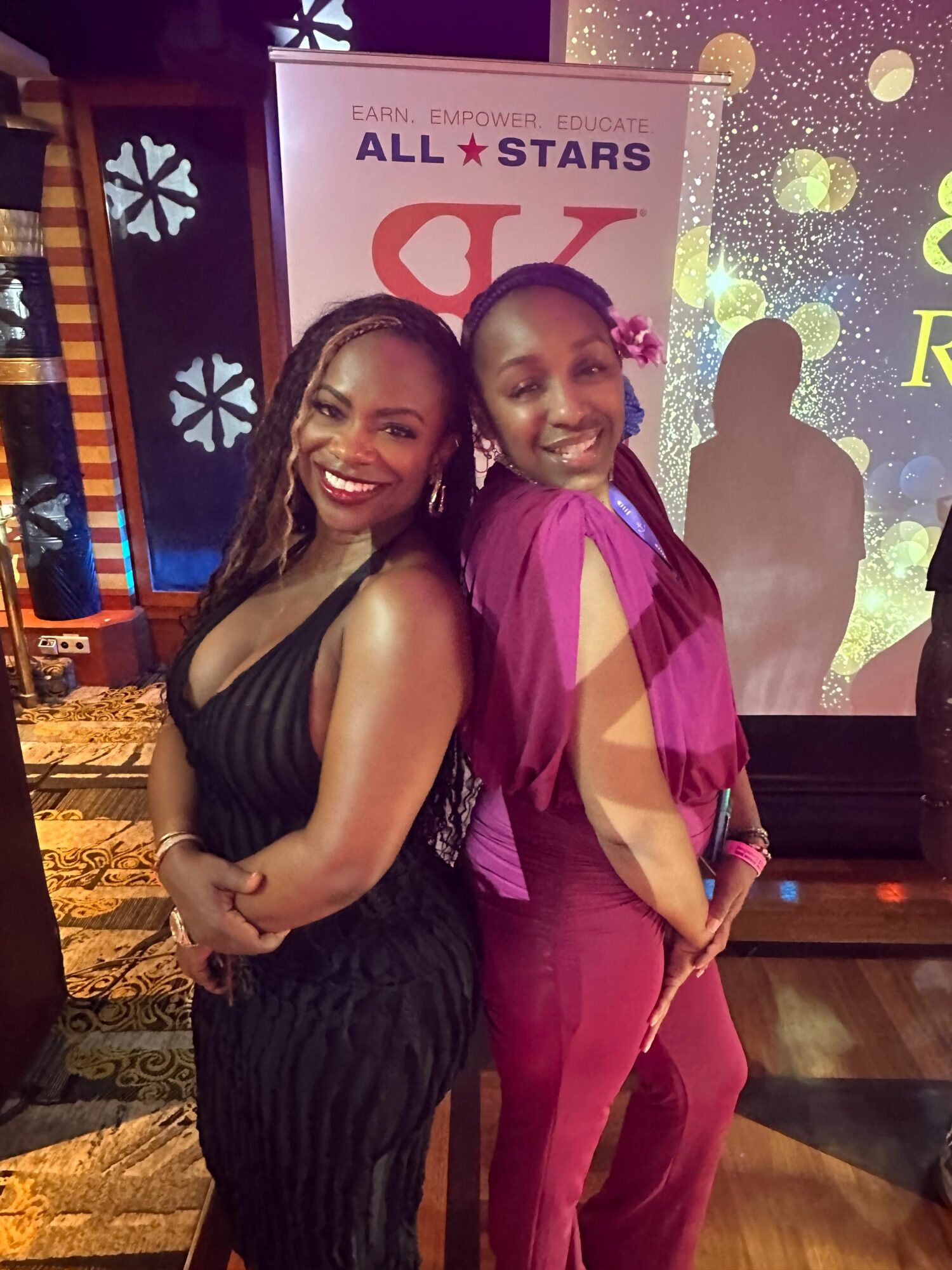 Two women posing together at an event, smiling, with a colorful background and a sign that reads 'ALL STARS'.