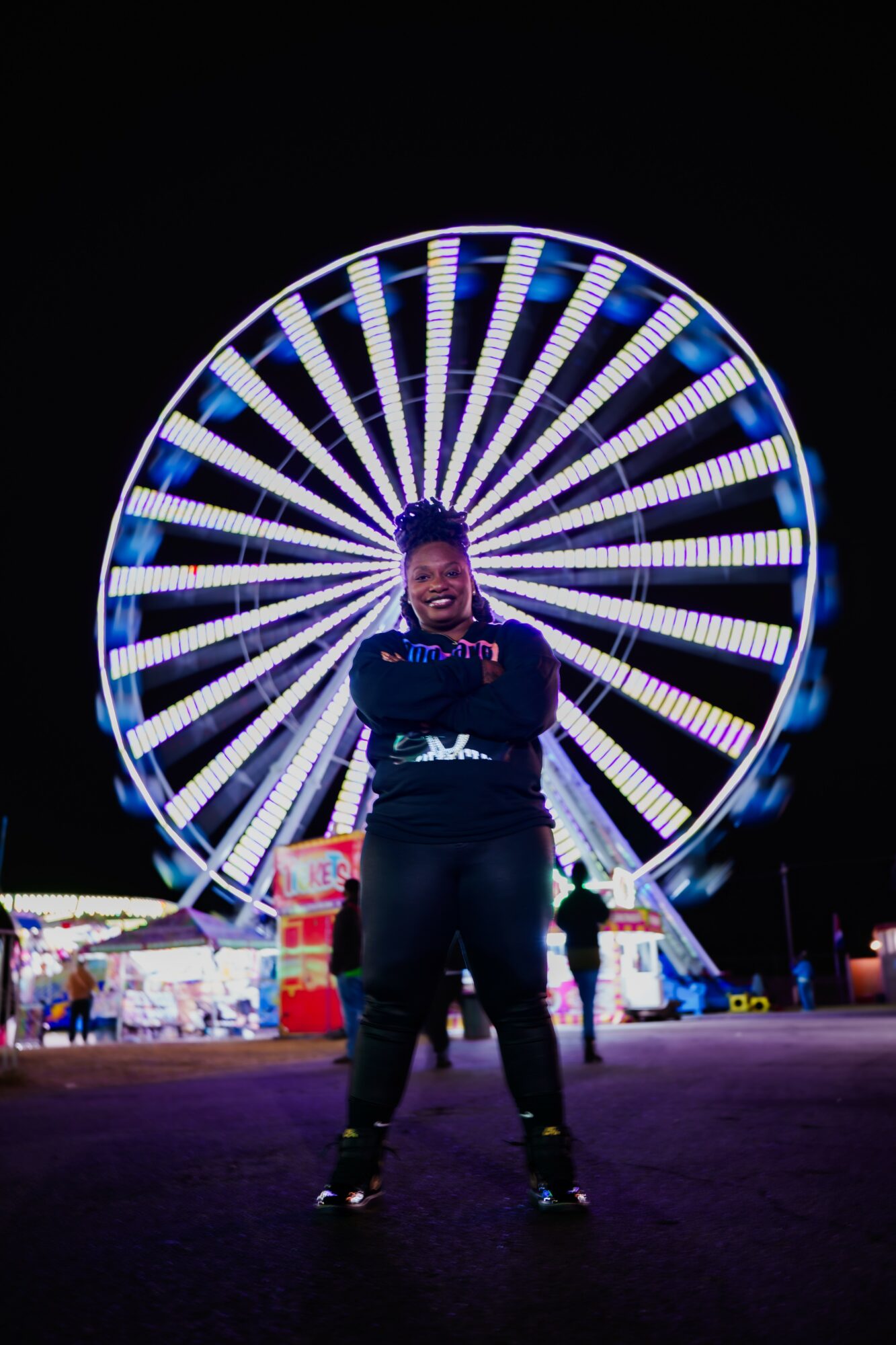 Person standing in front of a brightly lit Ferris wheel at night, arms crossed, smiling.